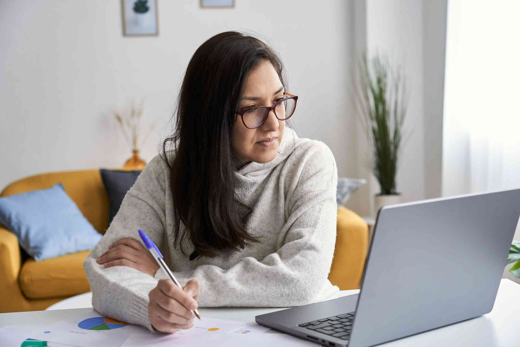 A woman in a white sweater sits at her desk with her laptop and writes on some papers spread out infront of her.