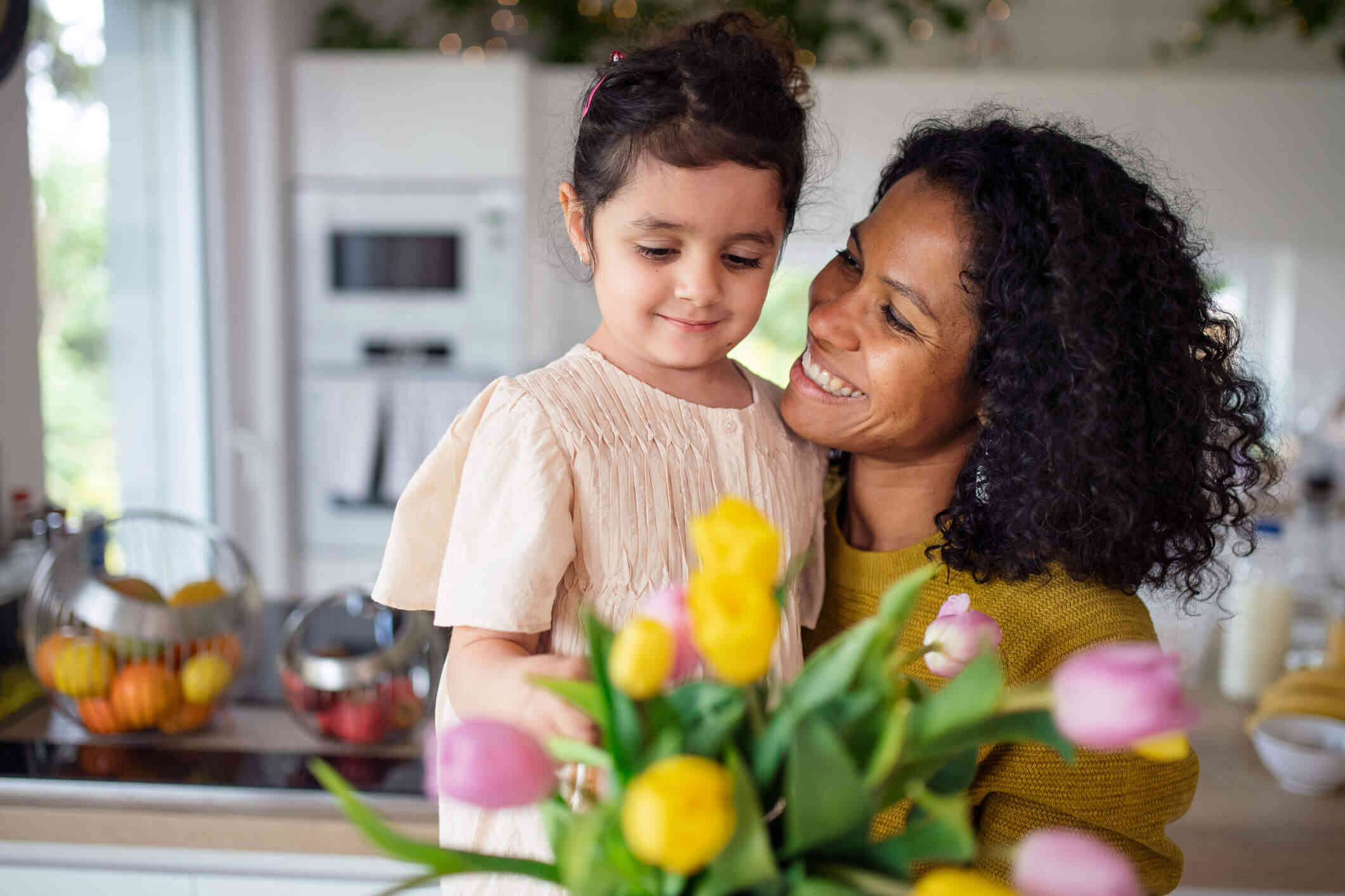 A woman in a yellow shirt holds her young daughter while smiling at her as they stand in their home near fresh cut flowers.