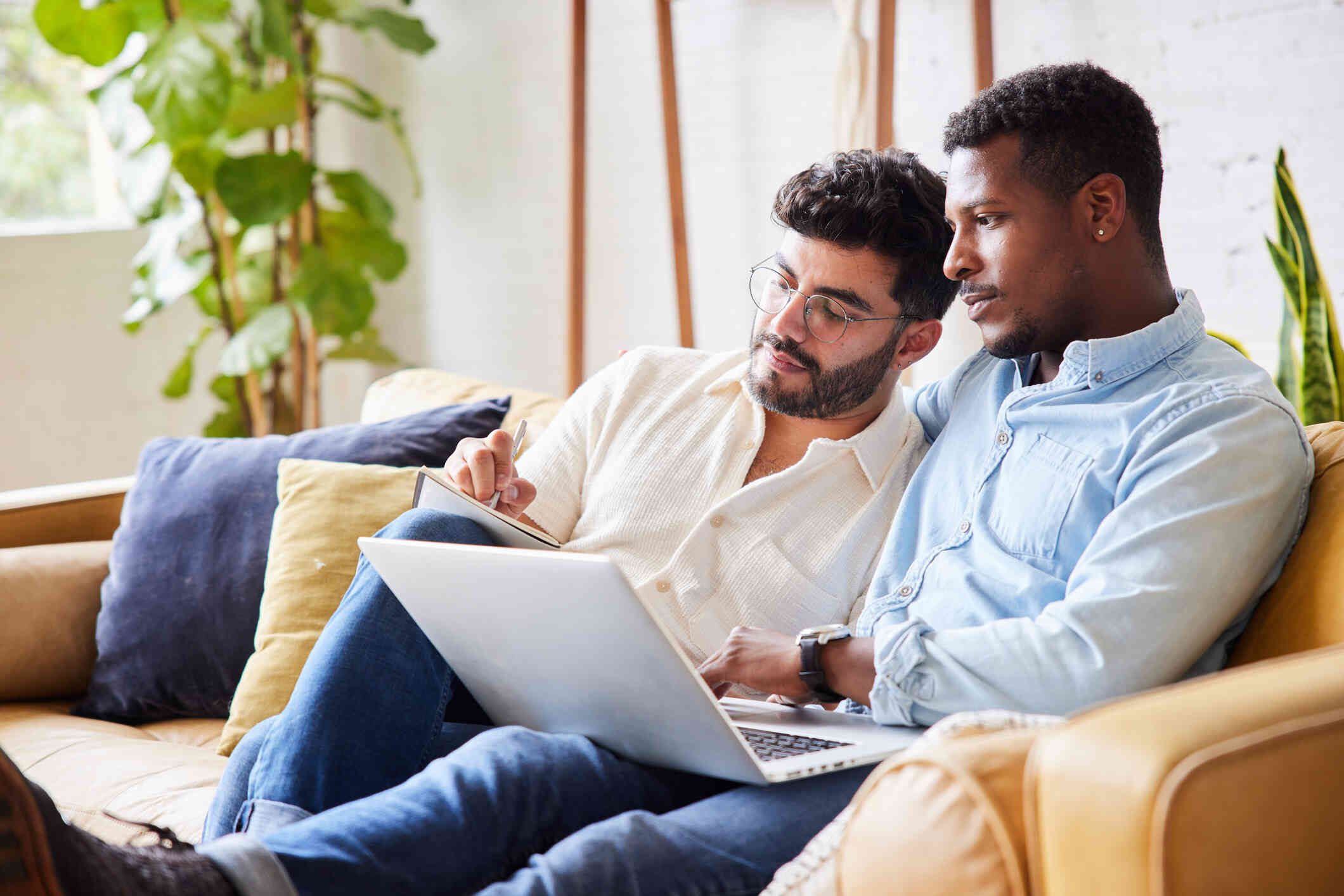 A man wearing a blue button up shirt sits on a couch with a laptop on his lap. Another man sits next to him wearing a white button up shirt and taking notes on a paper.