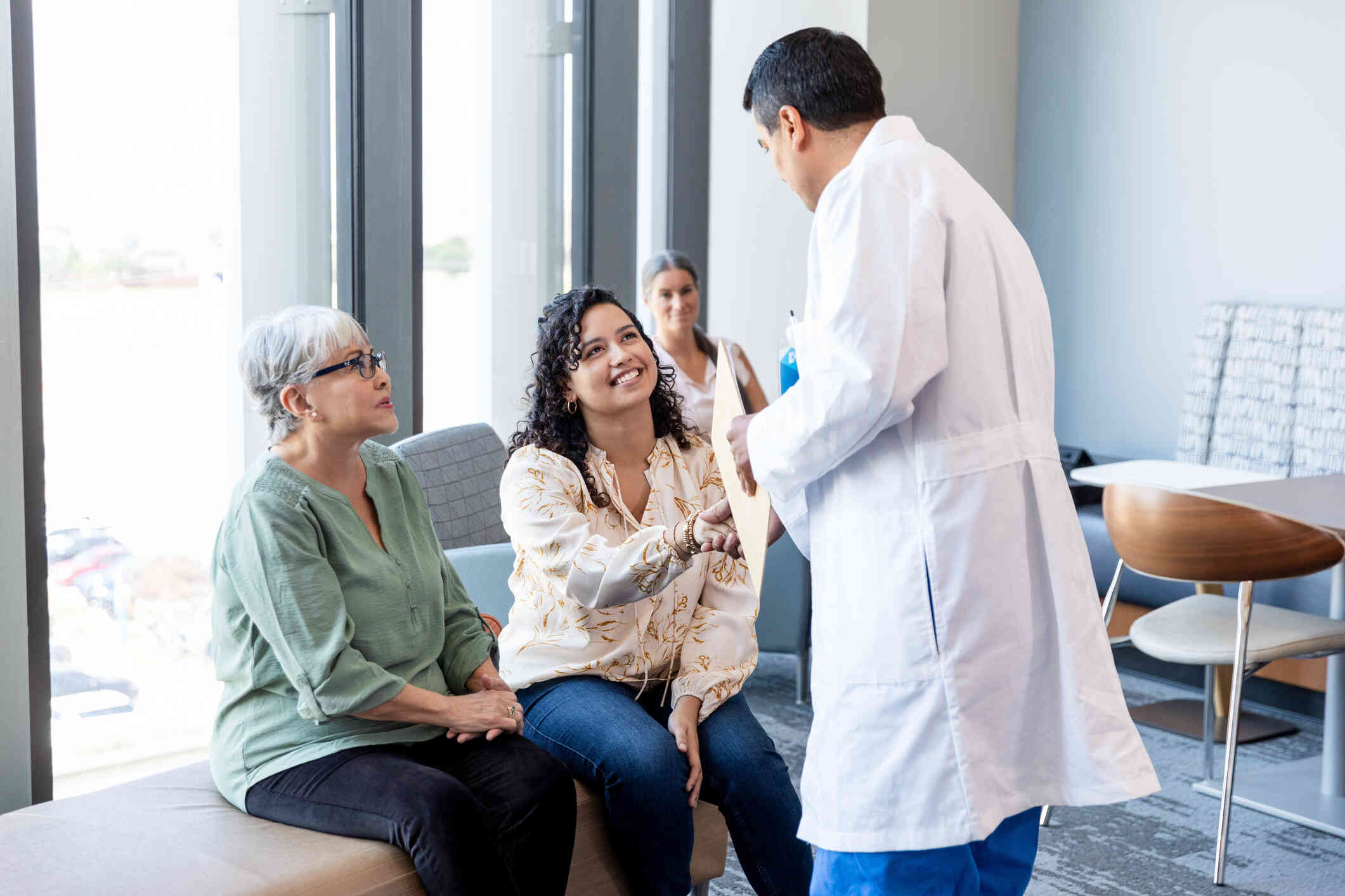 A woman with curly hair sits in a waiting room and smiles as she shakes the hand of a doctor in a white coat walking over to her. A mature woman sits in a chair next to her.