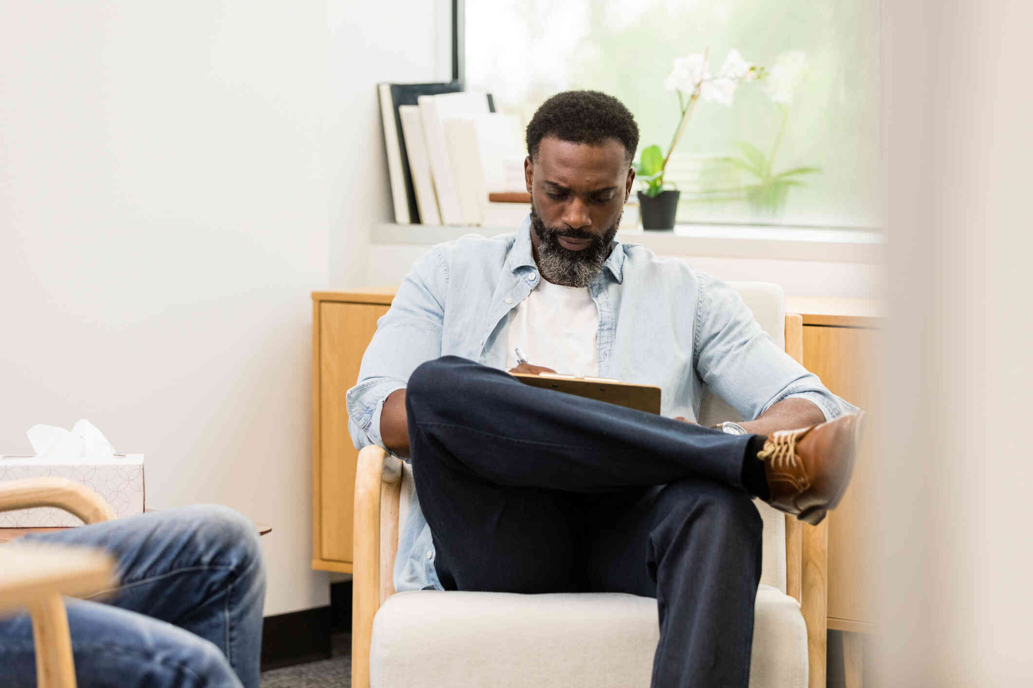 A close up of a male therapist as helsits in a chair and looks down at the clipboard in his lap while sitting near his male patient during a therapy session.