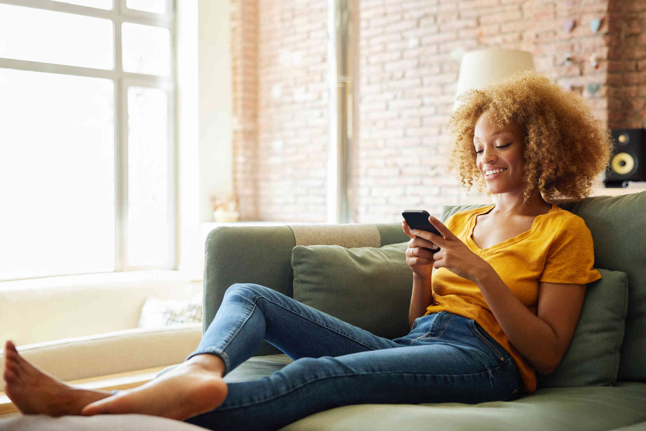 A woman in a yellow shirt sits on a green couch in her home and smiles down at the cellphone in her hands.