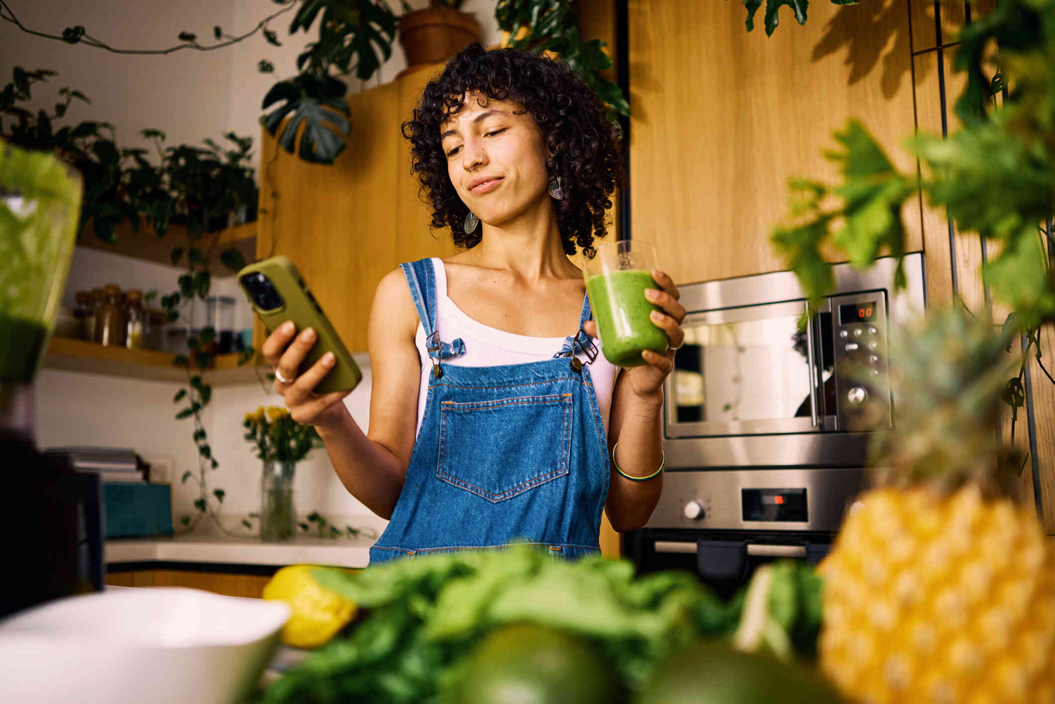 A woman in blue overalls stands in her kitchen and looks down at the phone in her hand while holding a cup of green juice in her other hand.