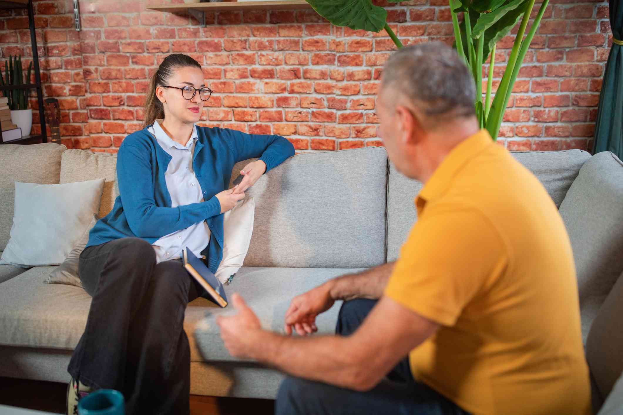 A woman ina blue cardigan sits on the couch across from her male therapist and listens as he talks during a therapy session.