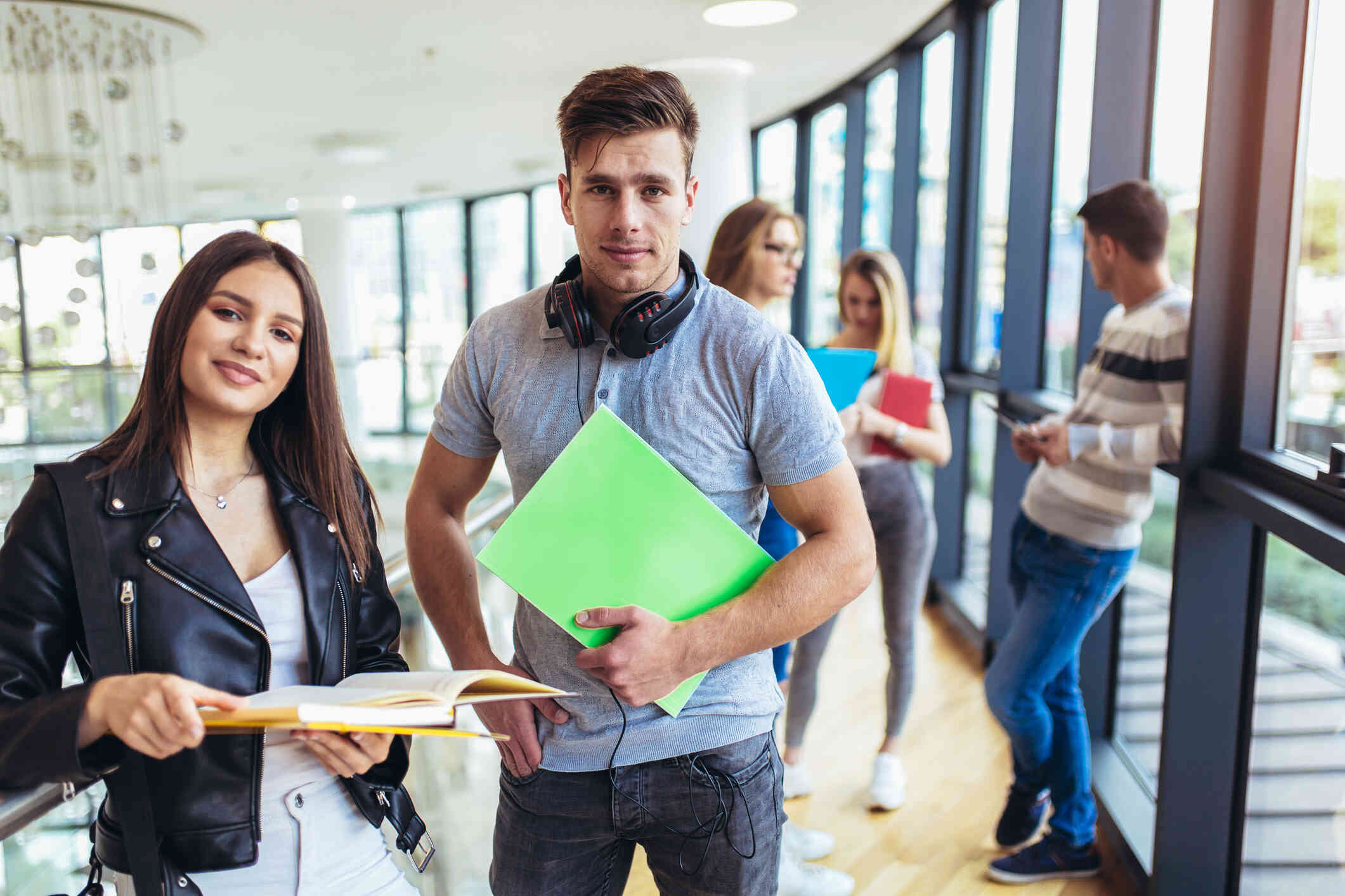 A male and female college student stand in a busy school hallway while holding books and looking at the camera.