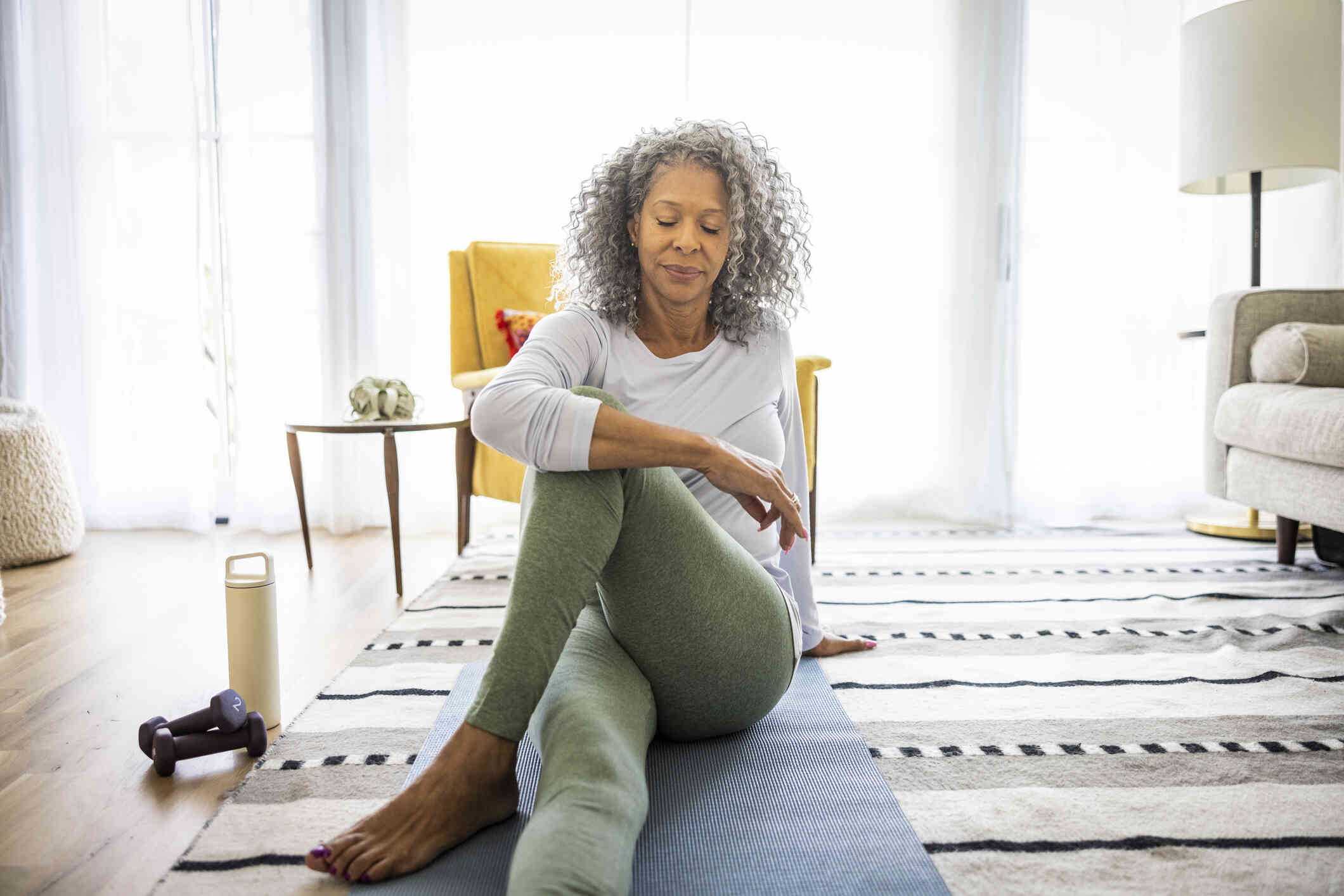 A middle aged woman sits on a yoga mat in her home and does theraputic stretches.