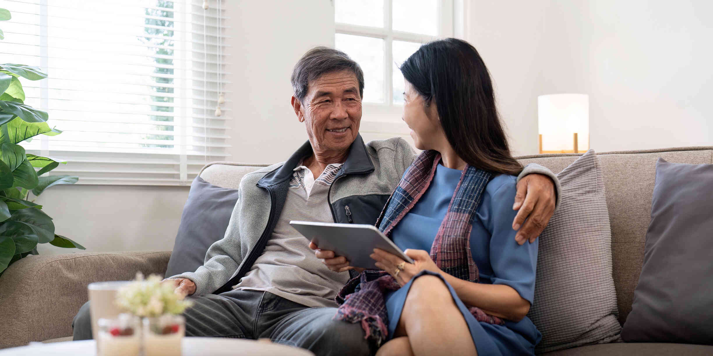 A mature male and female couple sit close together on the couch while smiling at one another.