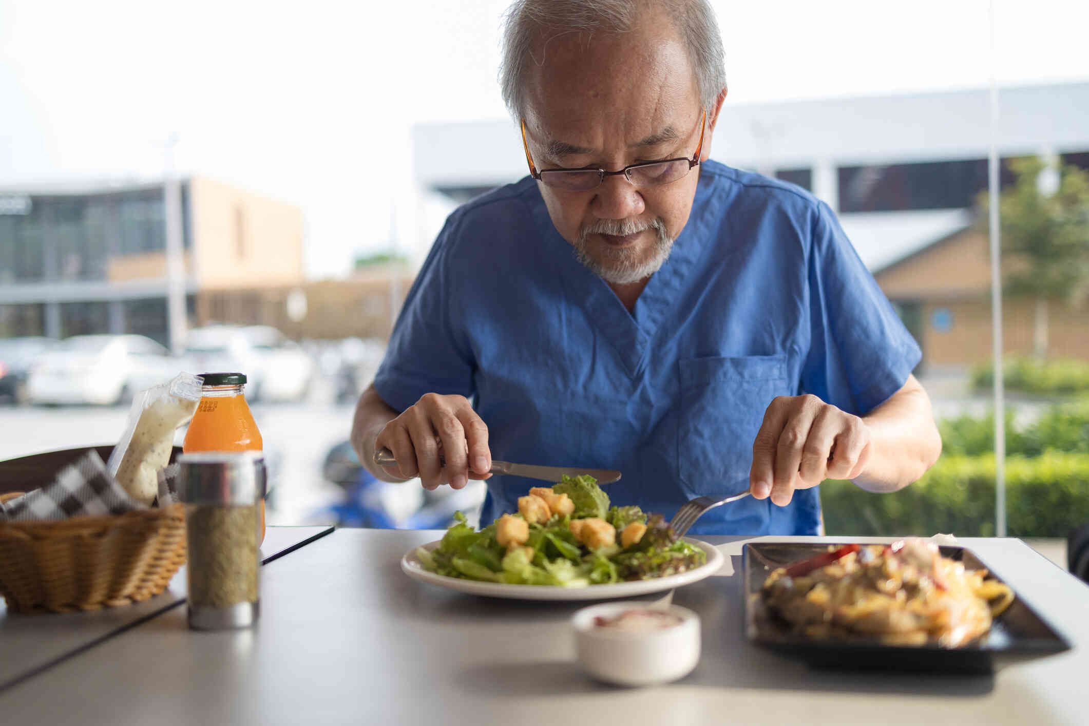 An elderly man in blue scrubs eats his salad at the table.