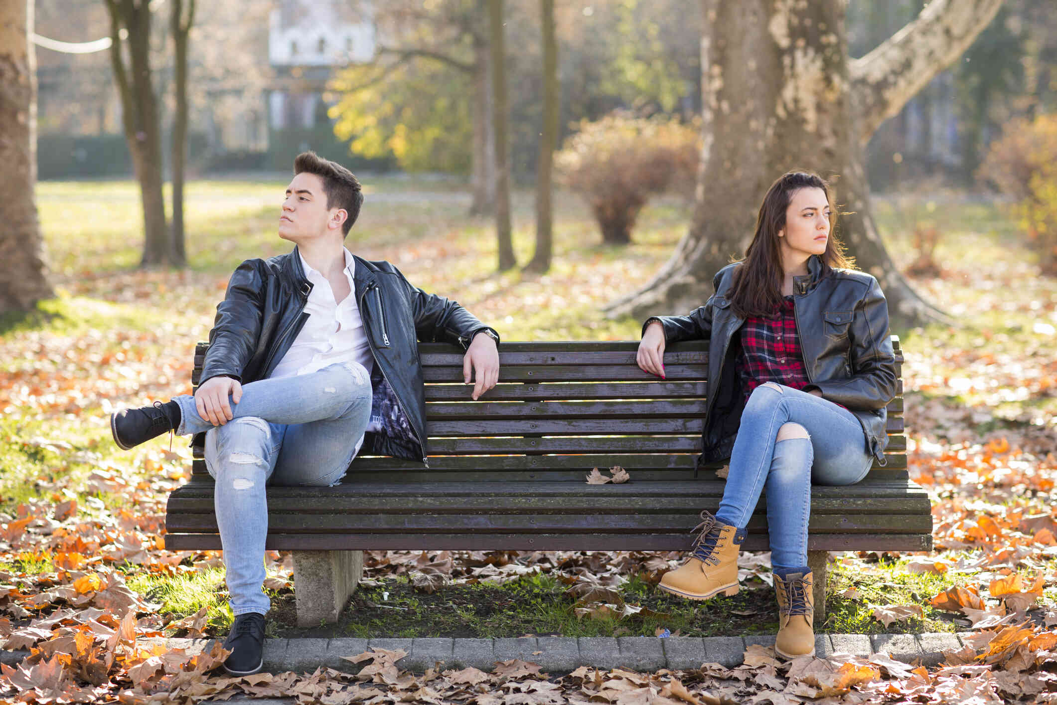 A male and female couple sit on opposite sides of a park bench on a sunny fall day while angrily looking away from each other.