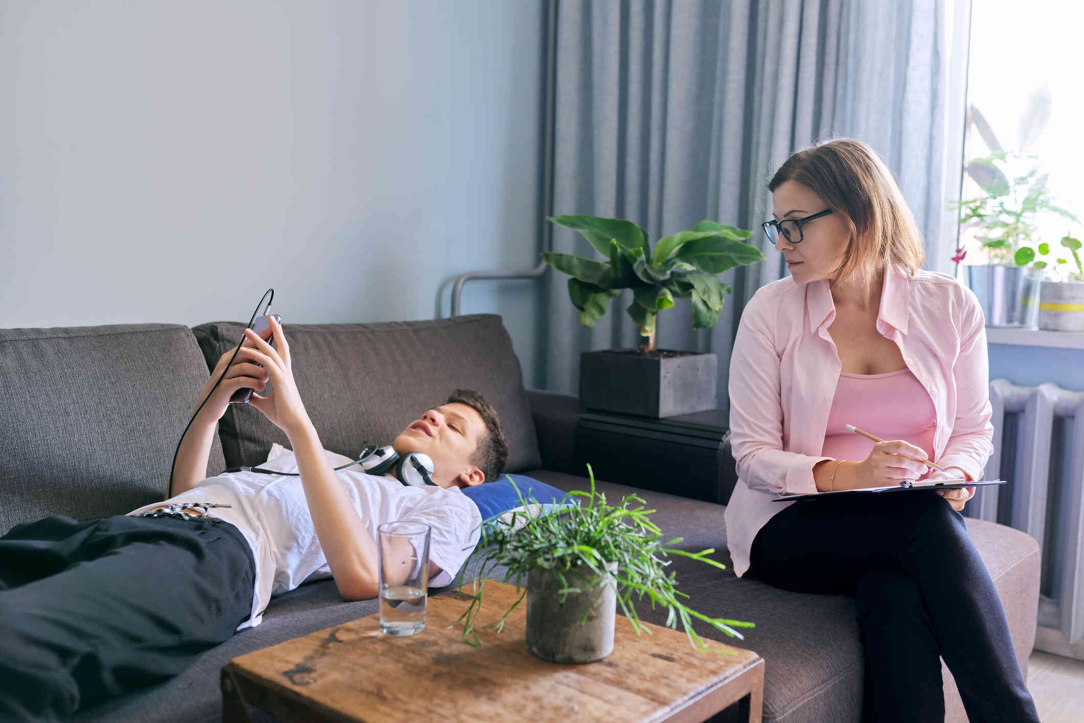 A teenage boy in a white shirt lays on the couch while looking at his phone as a young woman in a pink shirt sits across from him holding a clipboard