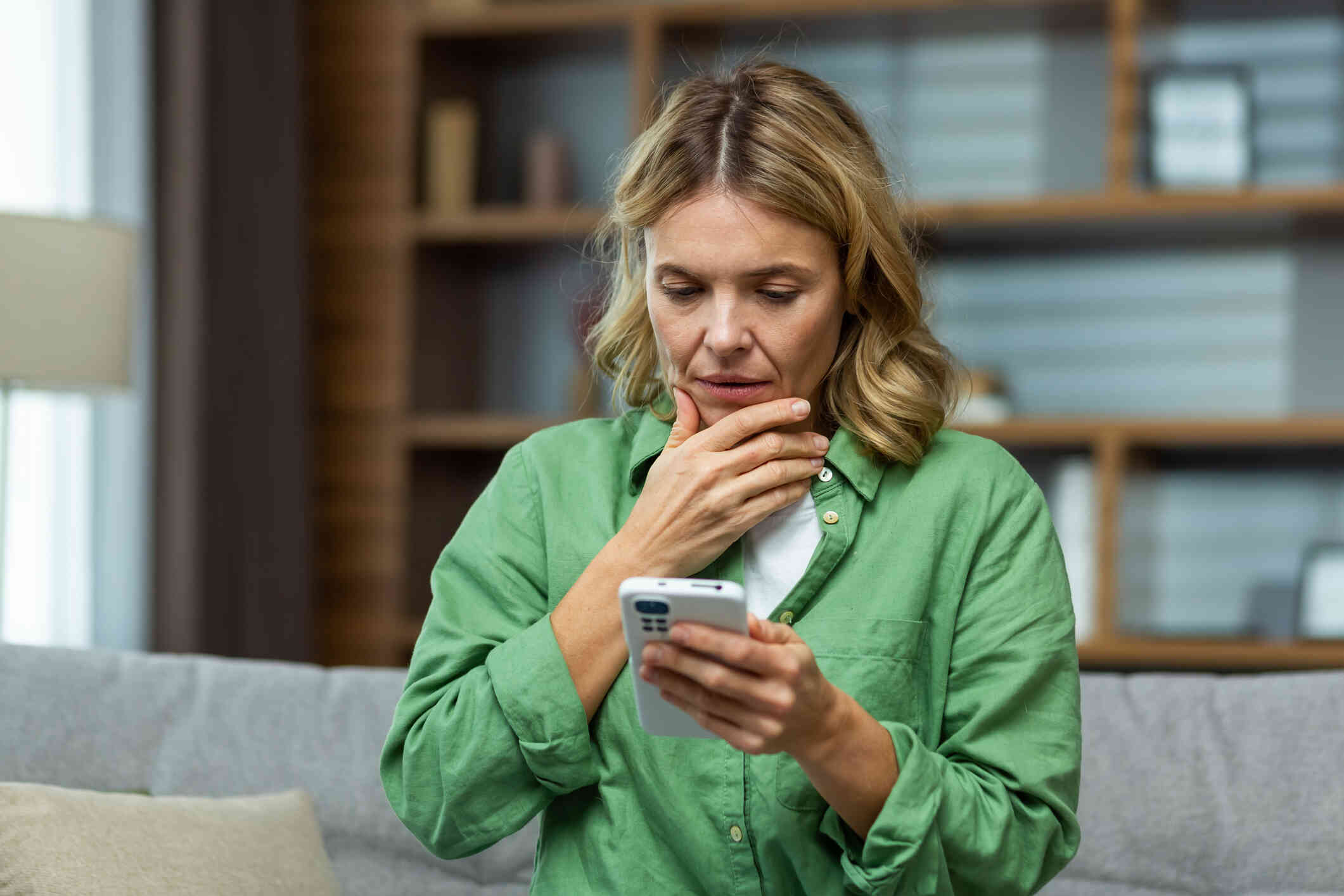 A woman in a green shirt sits on her home and looks down at the cellphone in her hand with a worried expression.