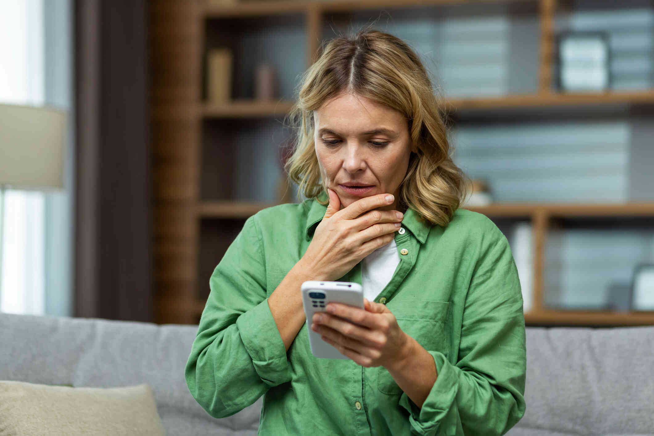 A woman in a green jacket looks down at her phone with a shocked expression on her face.