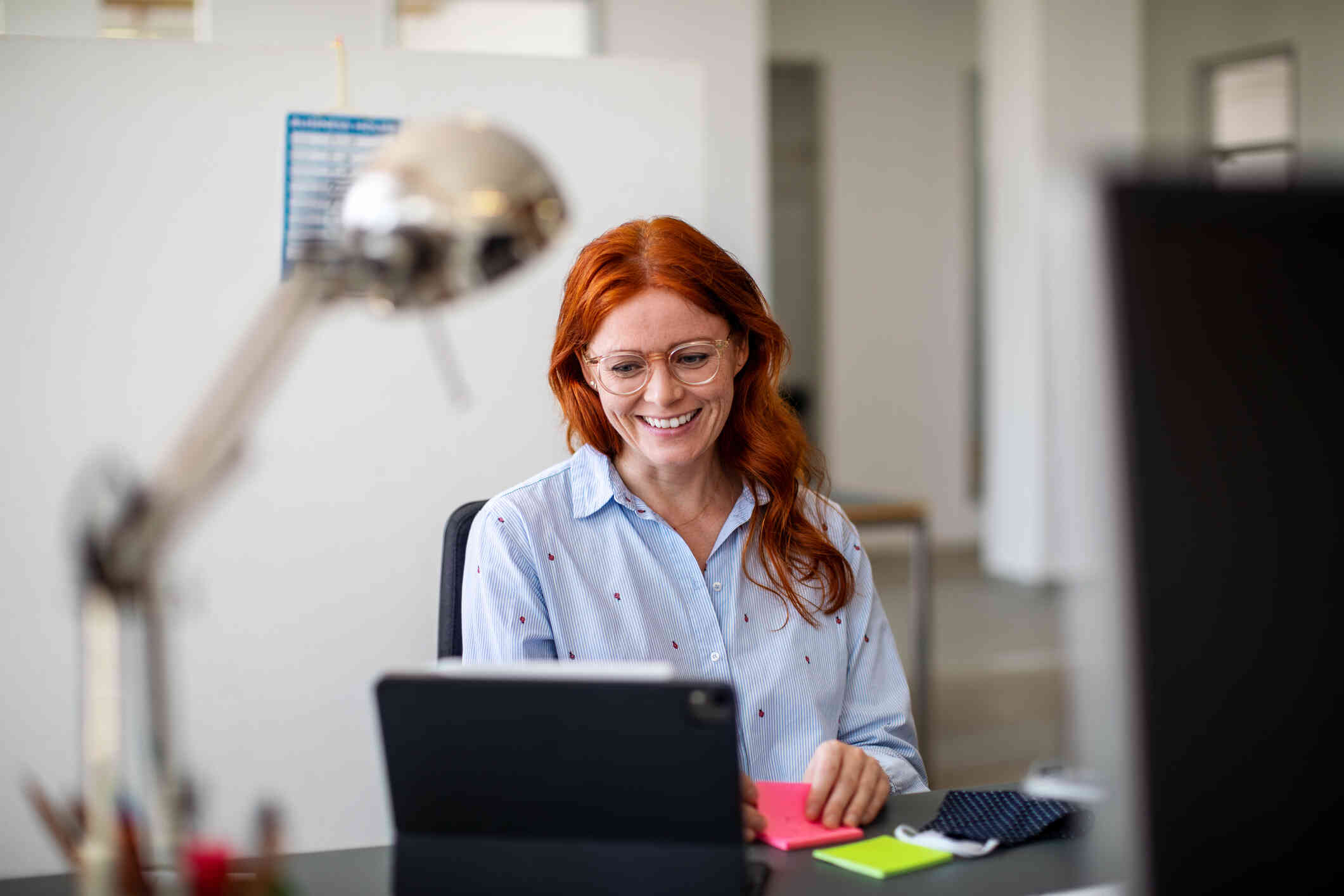 A woman seated at a table smiles while looking at her tablet screen, holding a sticky note. She appears to be on a video call.