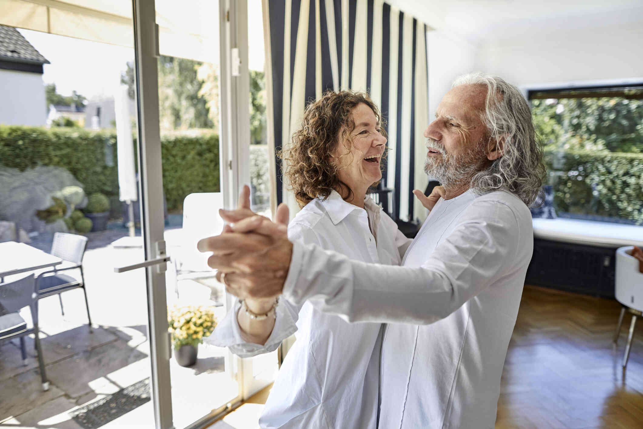 A mature man and woman both wear white shirts and smile as they hold each other and dance in their living room.