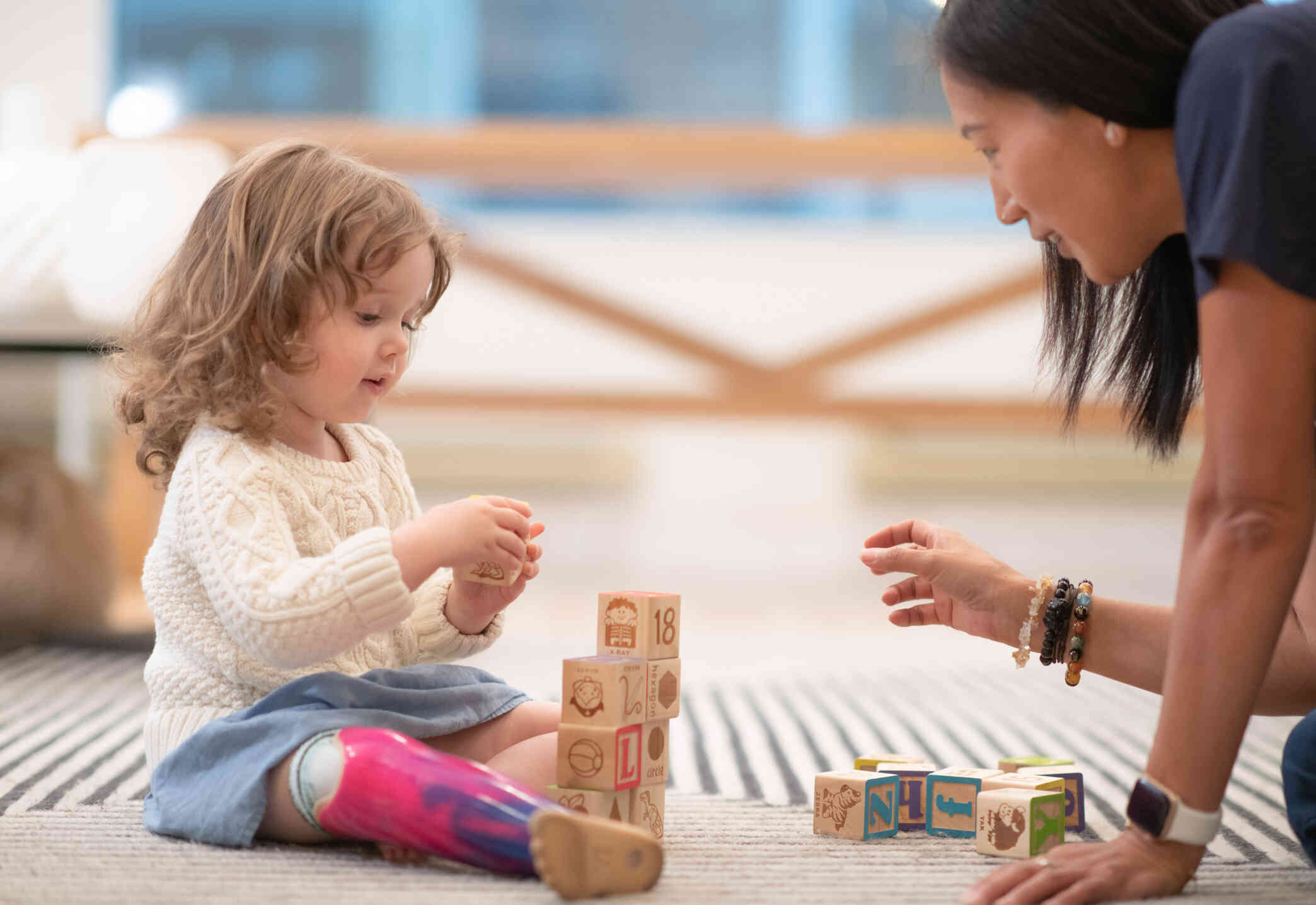 A young girl with a prosthetic leg sits on the foor and plays with some blocks as a woman leans down to smile at her.