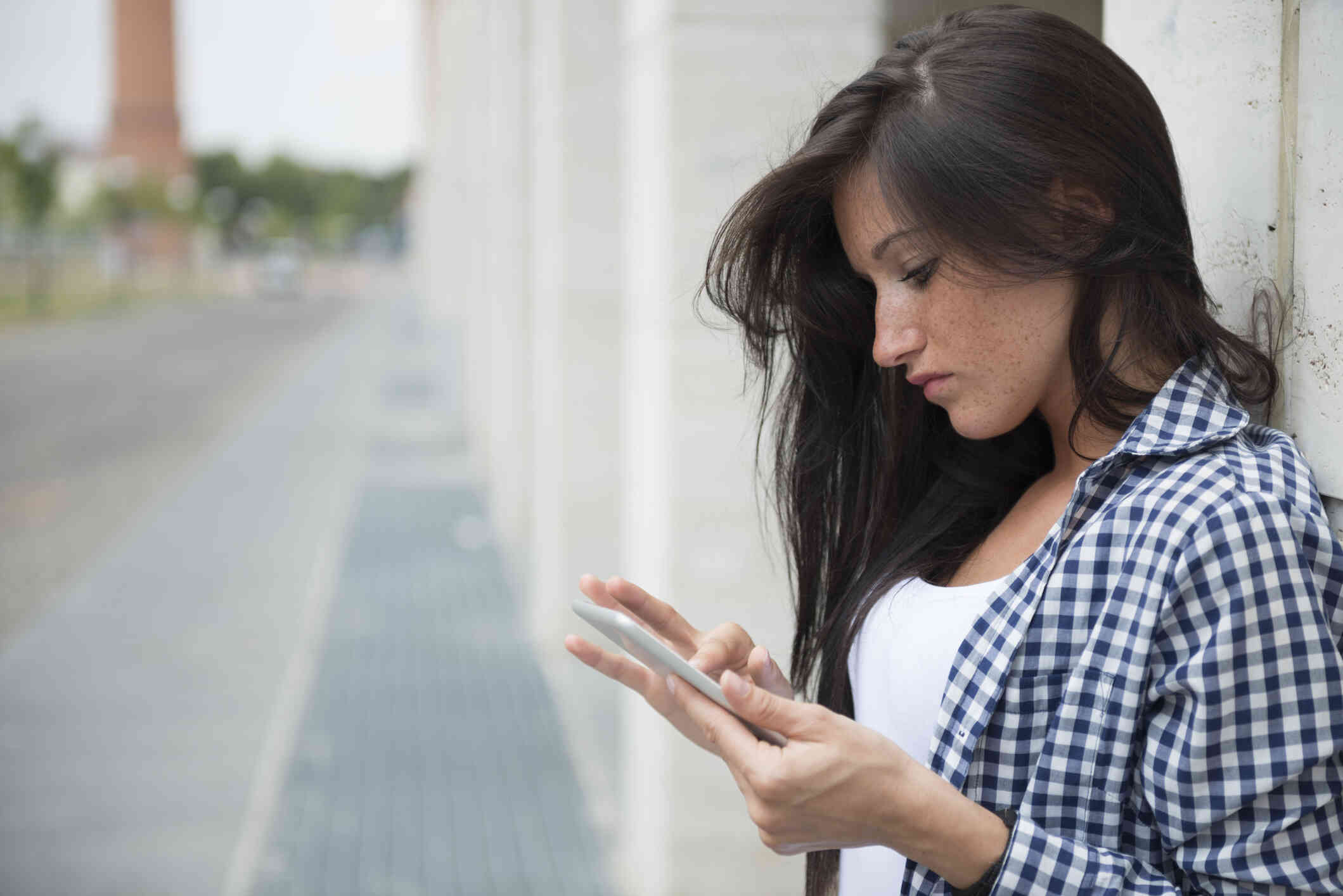 A woman in a plaid shirt leans against a wall while looking down at the phone in her hand.