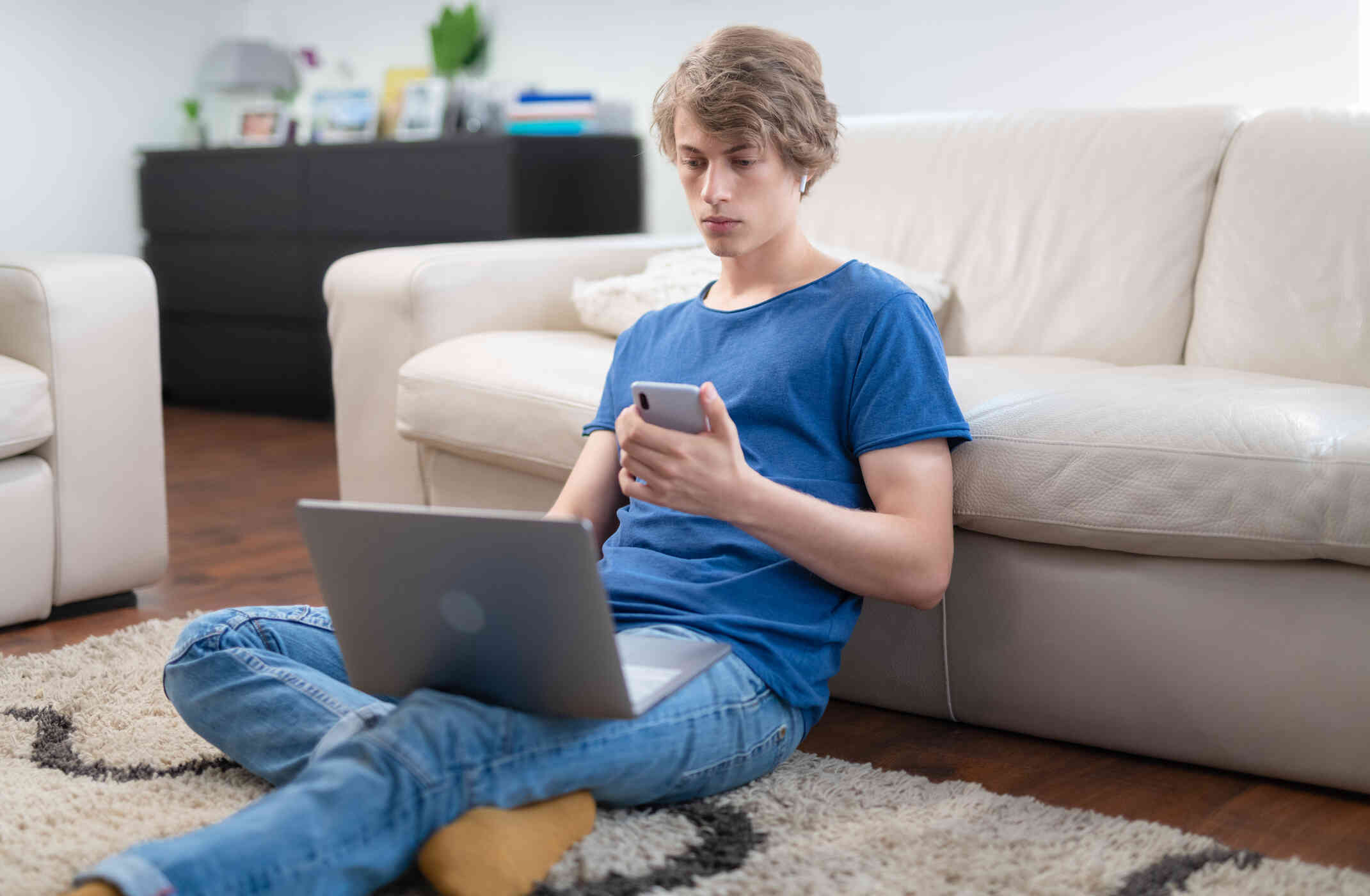 Ateen boy in a blie shirt sits on the florr with his back against the couch and his laptop open in his lap as he looks at the cellphone in his hand.
