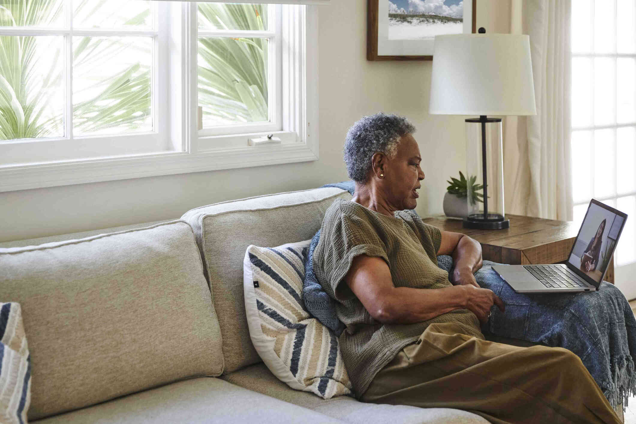 A middle aged woman in a green shirt sits on her couch and talks to the female therapist on the laptop that is open infront of her.