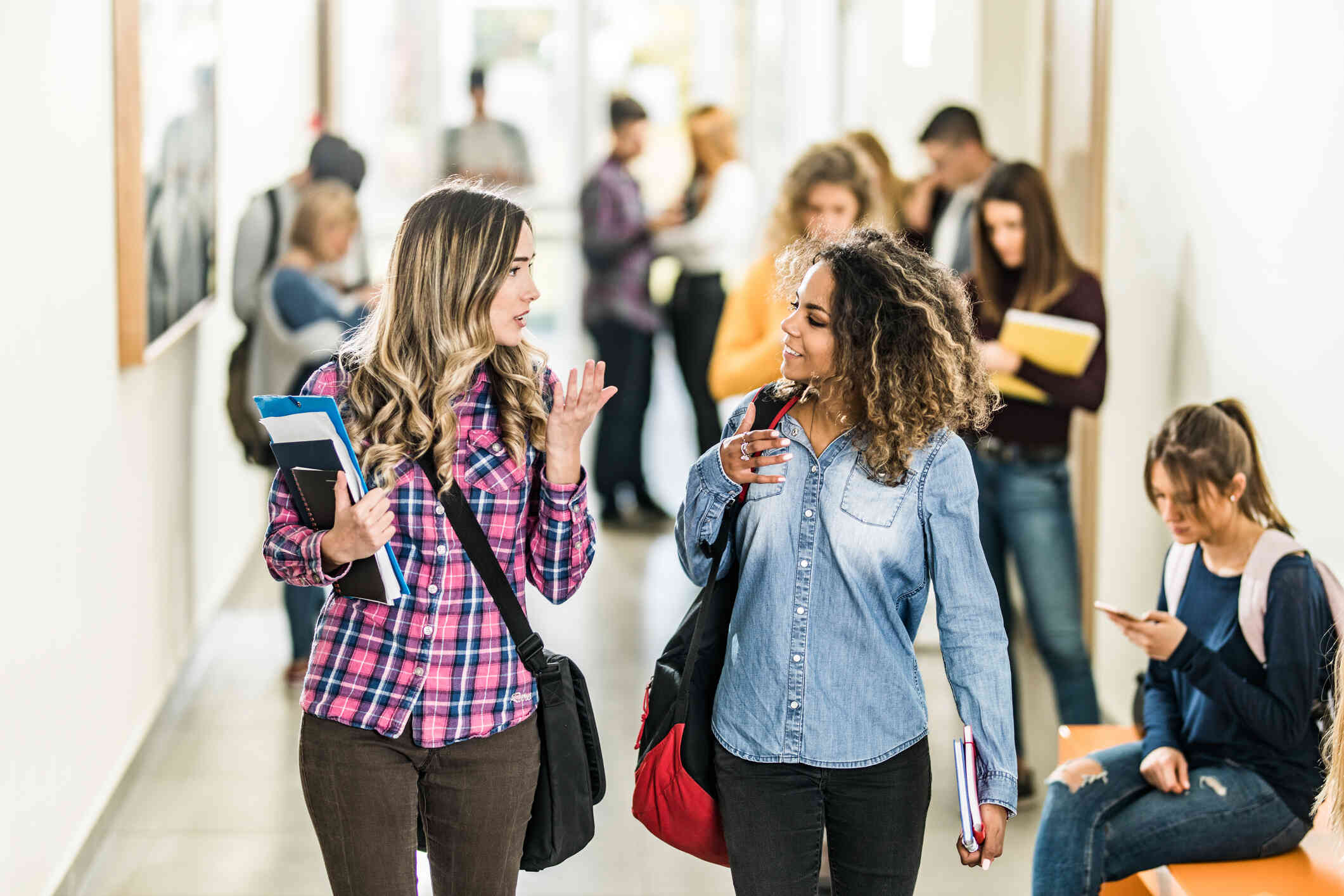 Two young women walk down a crowded hall with backpacks and books. One woman with a pink plaid shirt is speaking while the other woman in a denim shirt smiles and listens. 