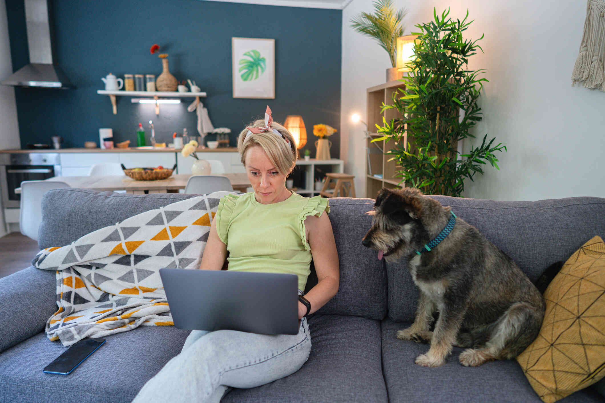 A woman ina green shirt sits on her couch with her grey dog next to her and types on the laptop open in her lap.