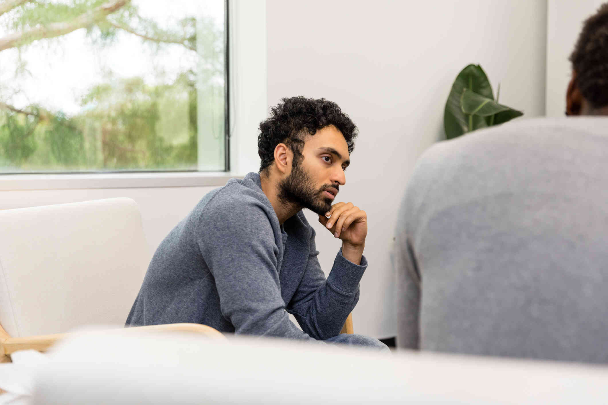 A man in a long sleeve grey shirt sits in a chair hunched over while listening to the man sitting across from him.