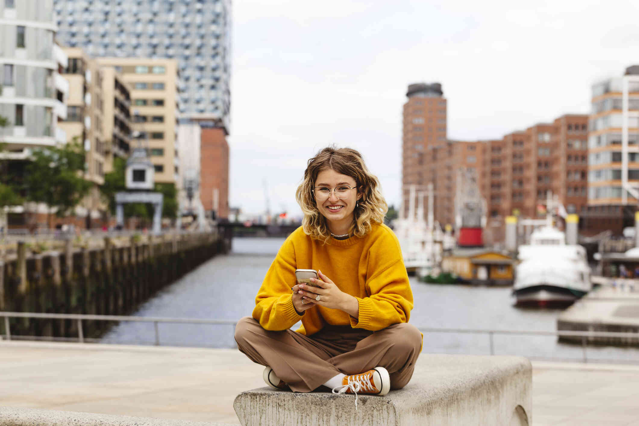 A woman sits cross legged at the dock while hilding her phone in her hand and smiling at the camera.