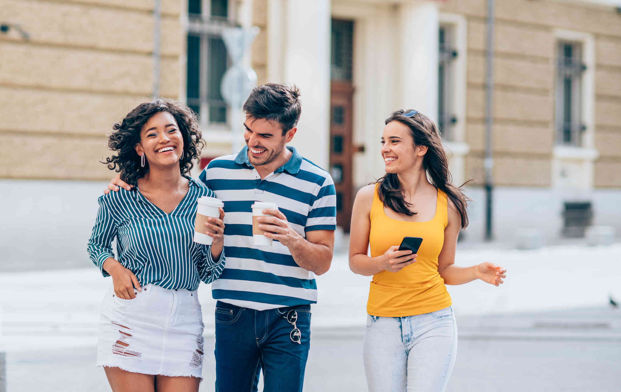 Three adult friends walk side by side outside on sunny day while smiling and laughing.