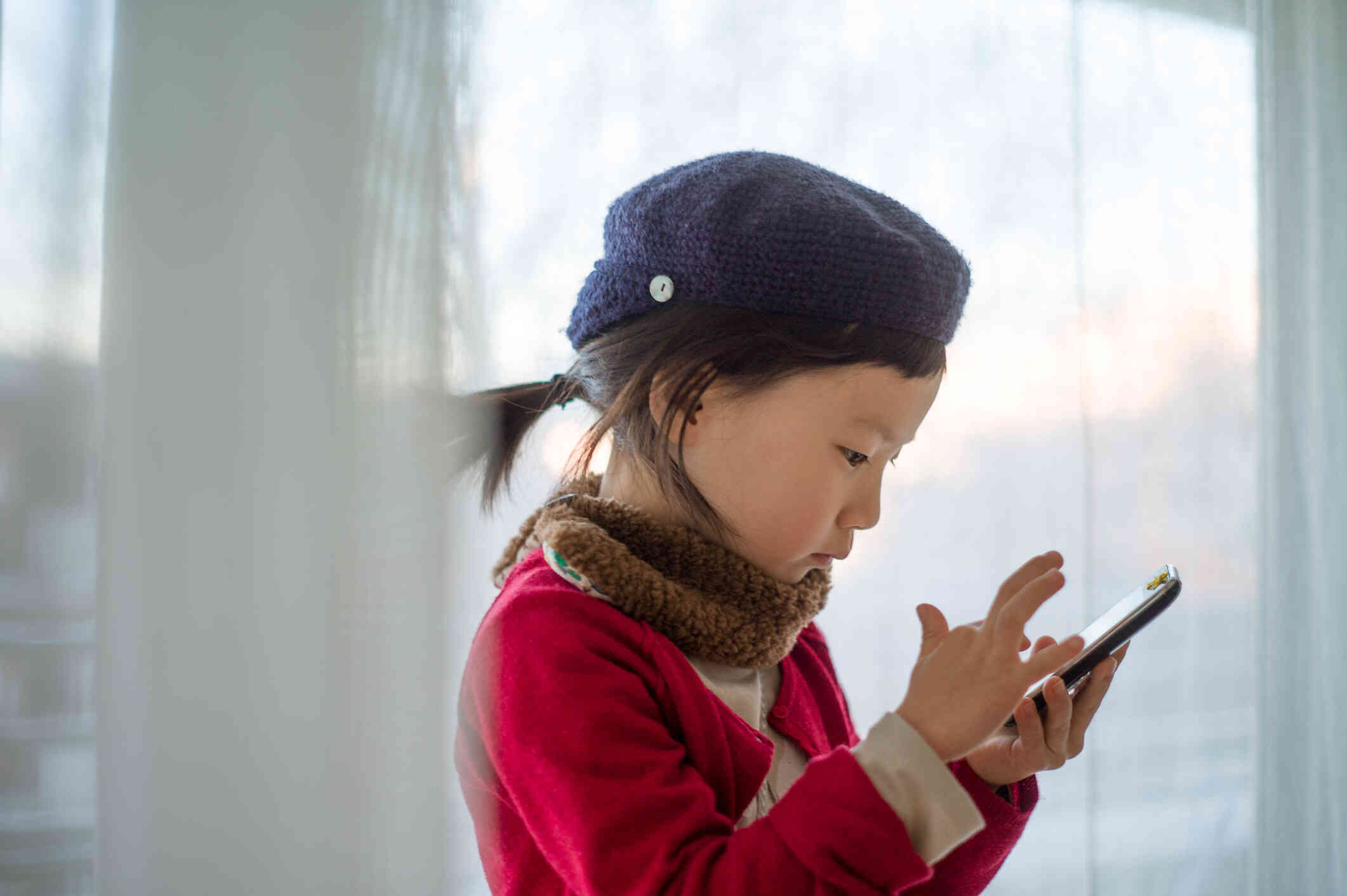 A young girl sits in her home and taps on the phone in her hand.