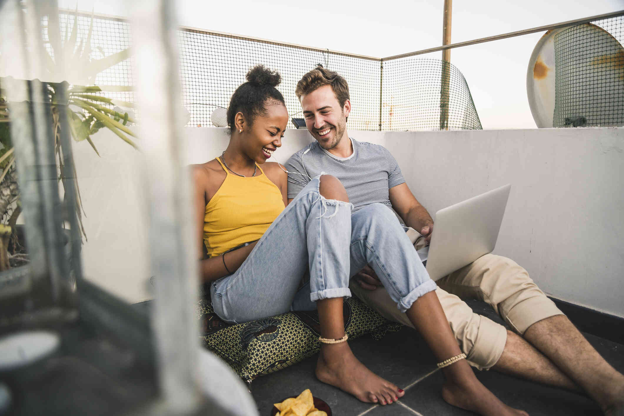 A man and woman sit side by side on the floor of a balcony and smile while sharing earbuds that are plugged into the laptop on the mans lap.