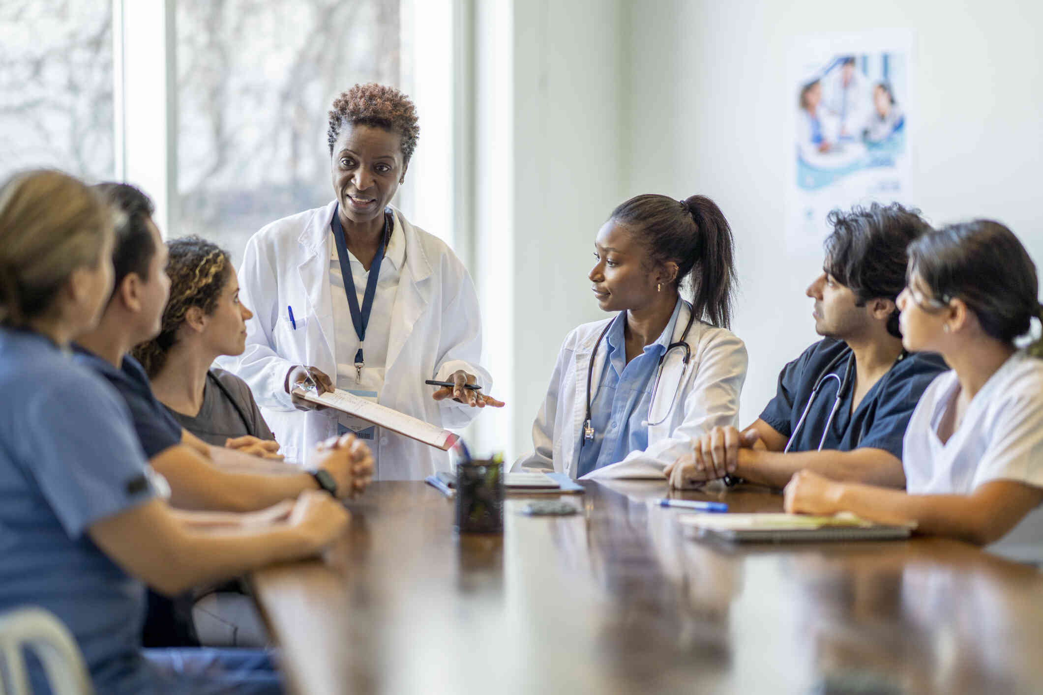 A woman in a white coat stands at the head of a table holding a clipboard and speaks with a group of male and female doctors sitting around the table with stethoscopes around their necks.