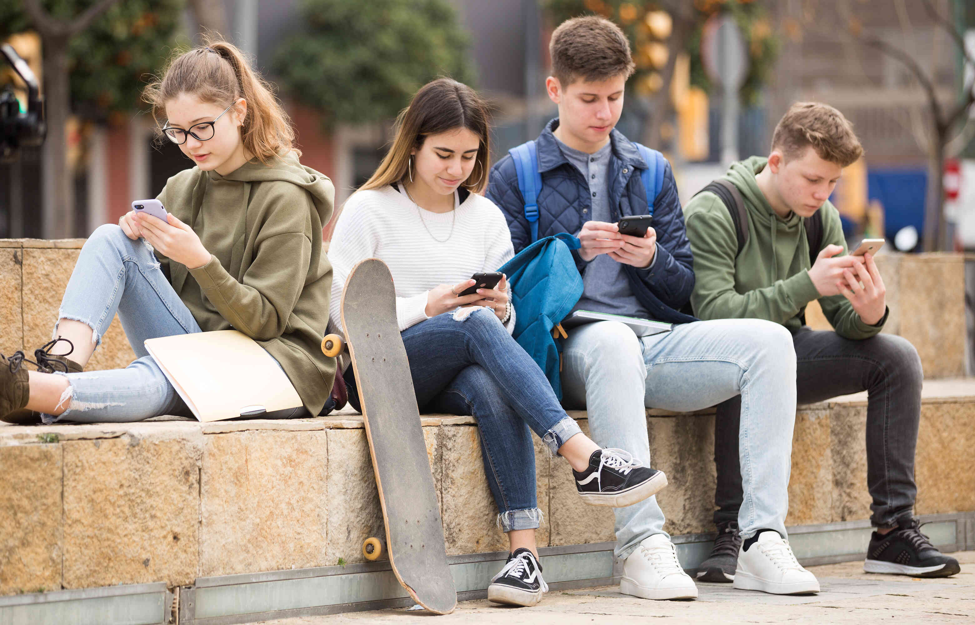 Four teenagers, two girls and two boys, sit outside on a bench looking down on their phones. They have backpacks and a skateboard.