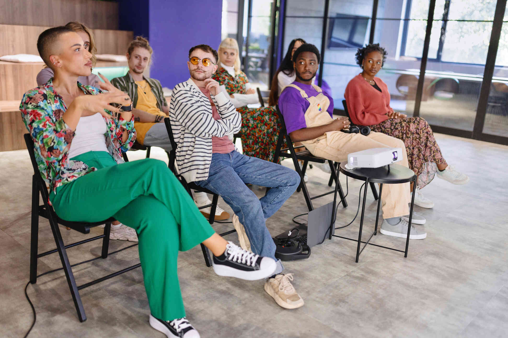 A group of people sit on chairs in a room and look towards a person with short hair and green pants who is speaking.