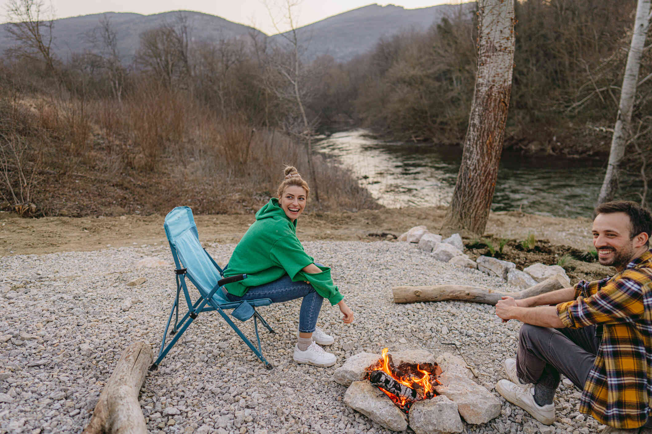 A woman wearing a green hoodie smiles as she sits outside in a camping chair next to a burning campfire. A man in plaid smiles as he sits across the fire from her.