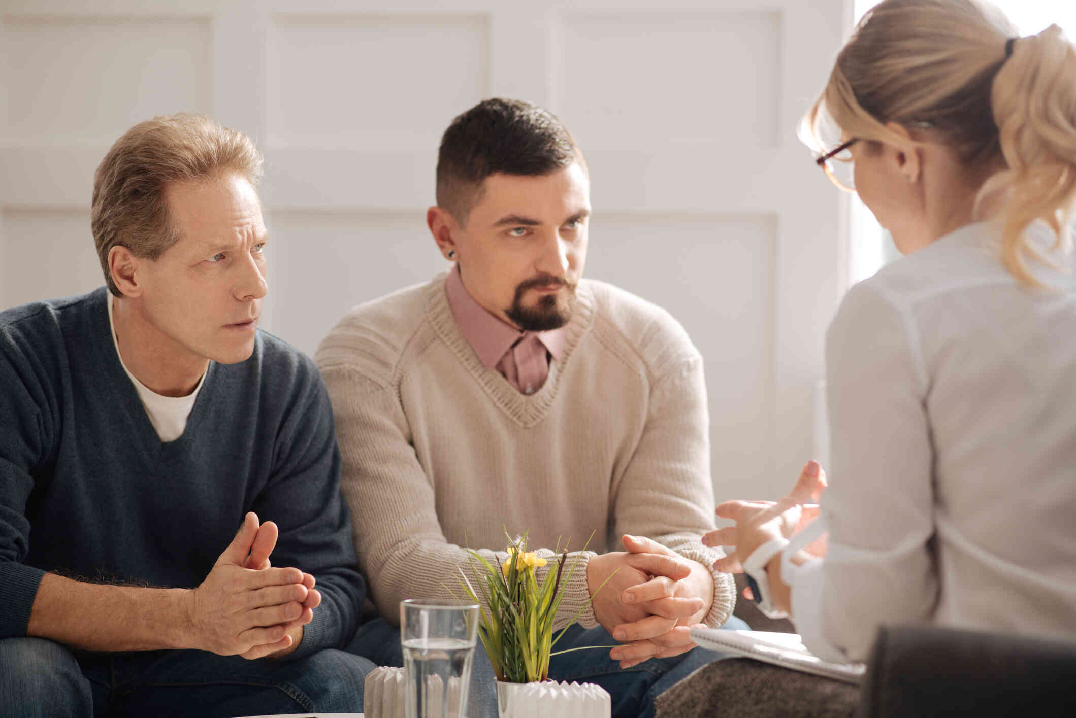A middle aged male couple sit across from their female therapist with serious expressions during a couples therapy session.
