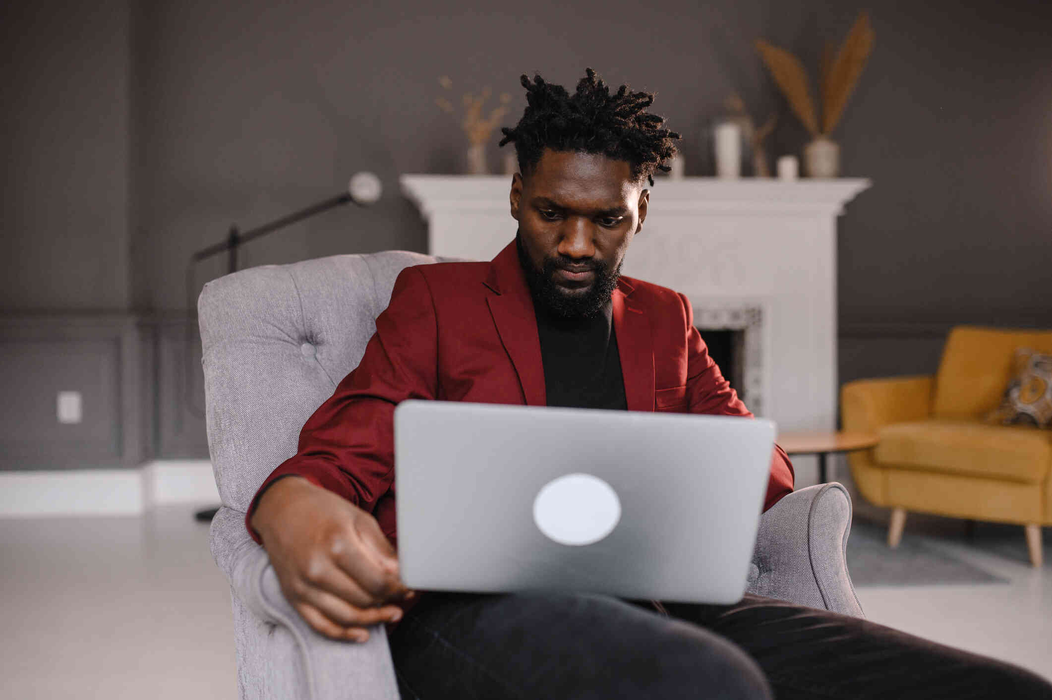 A man in a red blazer sits with his laptop open infront of him and looks at the screen with a serious expression.