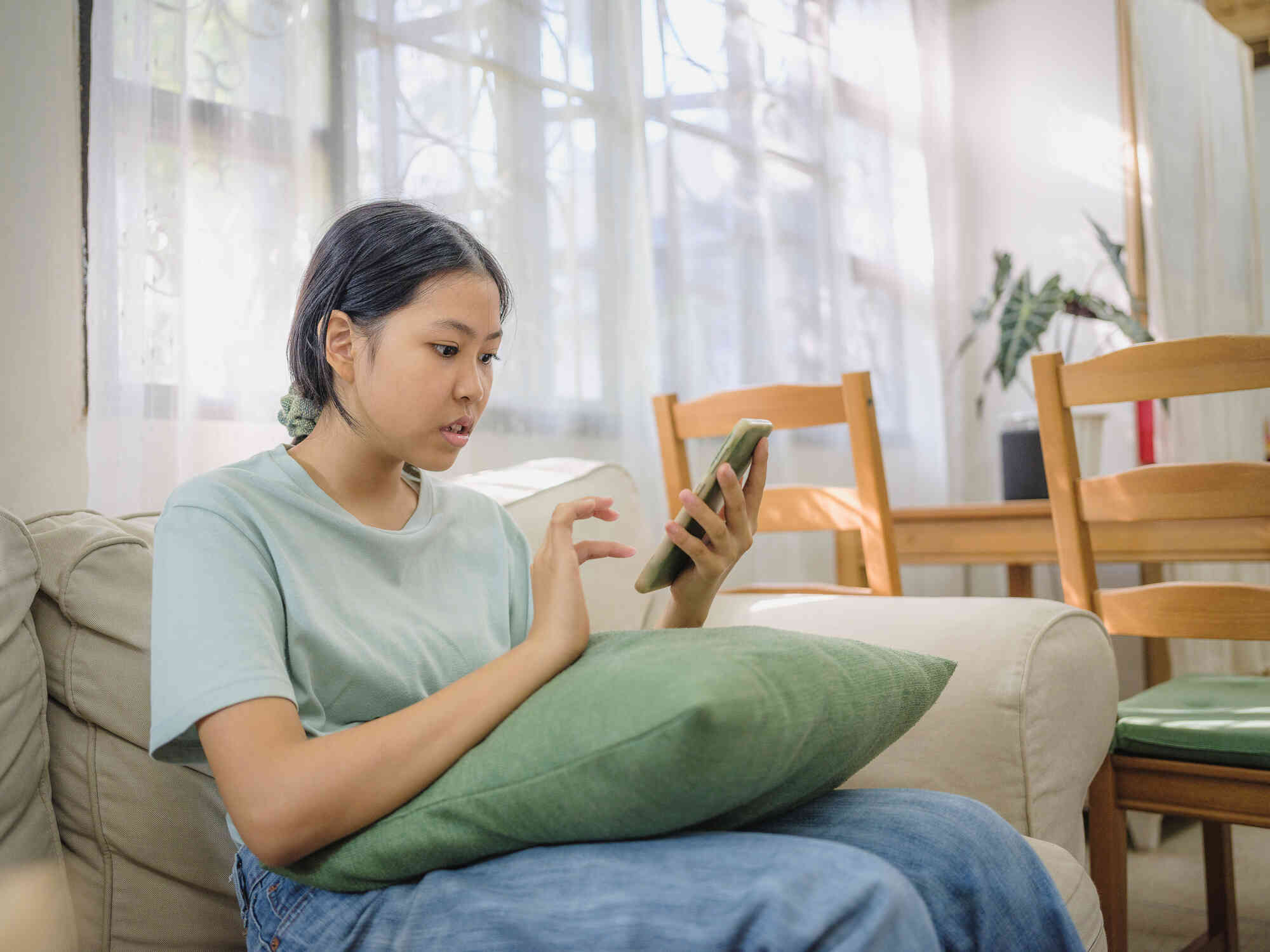 A teen girl in a green shirt sits on the couch and taps on her cellphone in her hand with a worried expression.