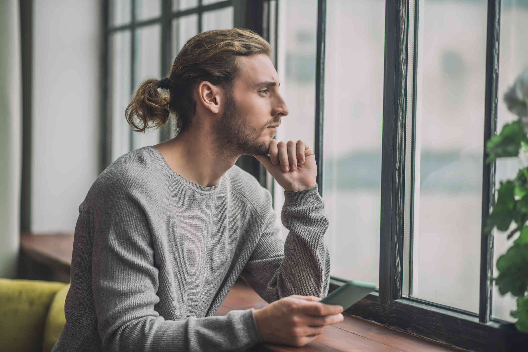 A man in a grey longsleeve shirt sits near a window with his phone in his hand as he gazes out sadly.