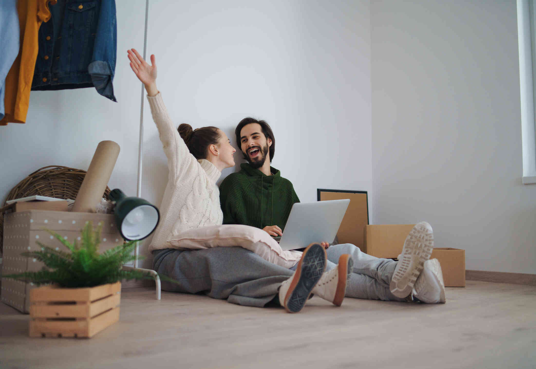 A woman in a white sweater raises her hand in the air as she smiles towards a man who smiles back and sits next to her on the floor with his laptop in his lap.