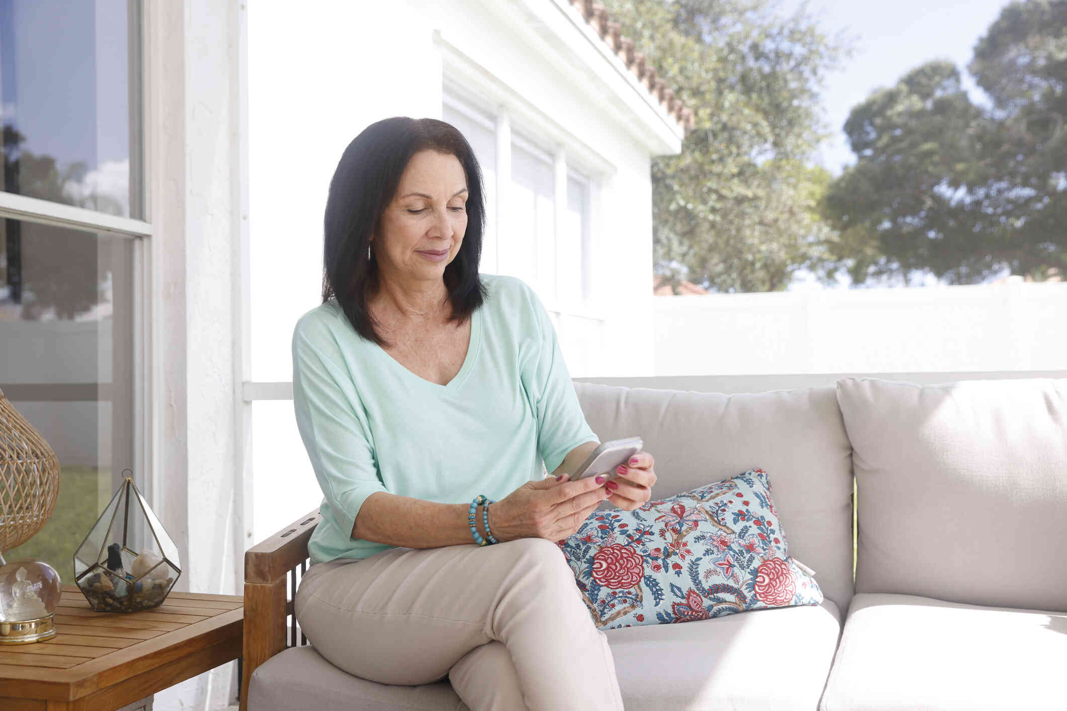 A woman in a green shirt sits on her couch in her home and loks down at the phone in her hands.