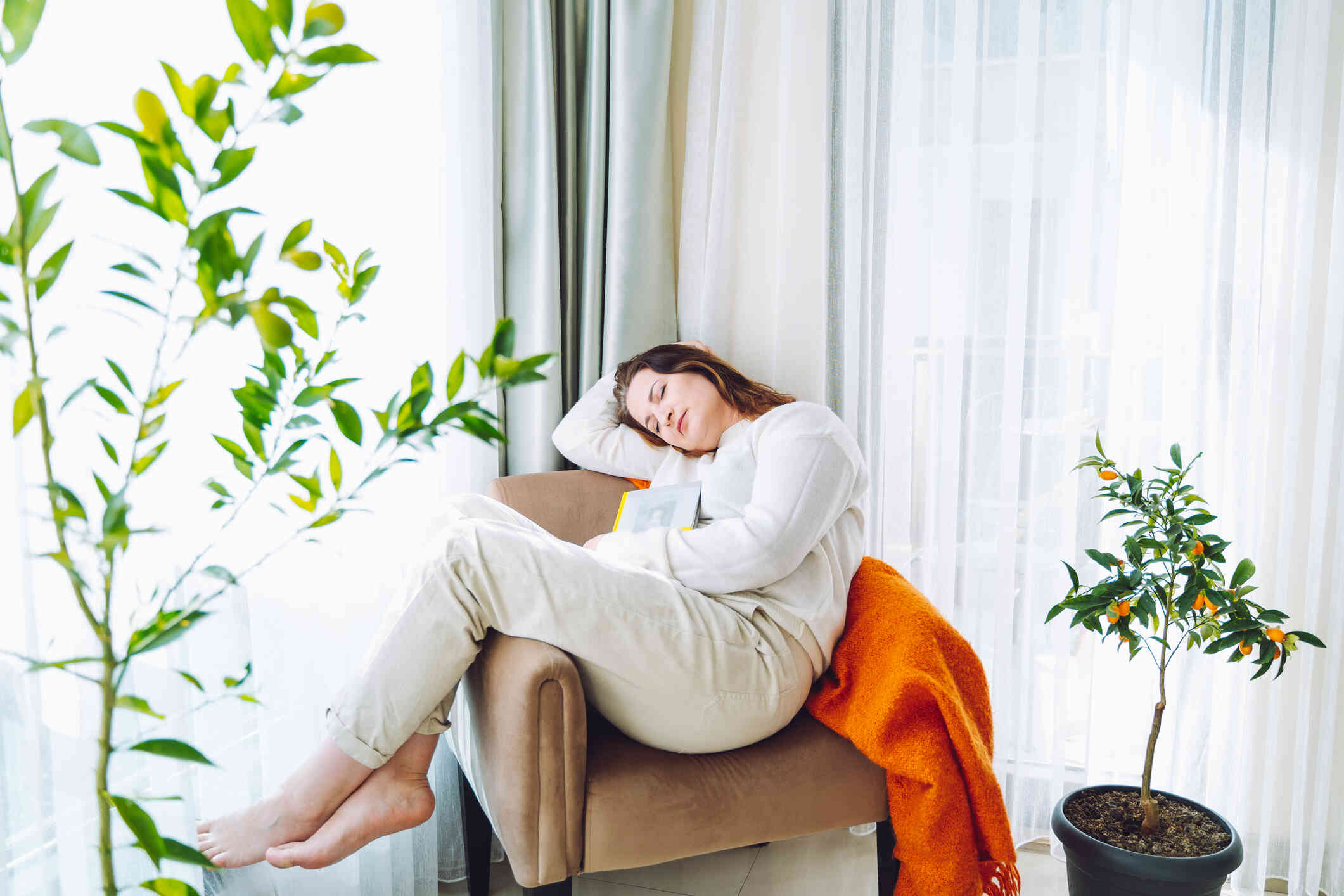 A woman in a white sweater sleeps in a chair with a book during the day.