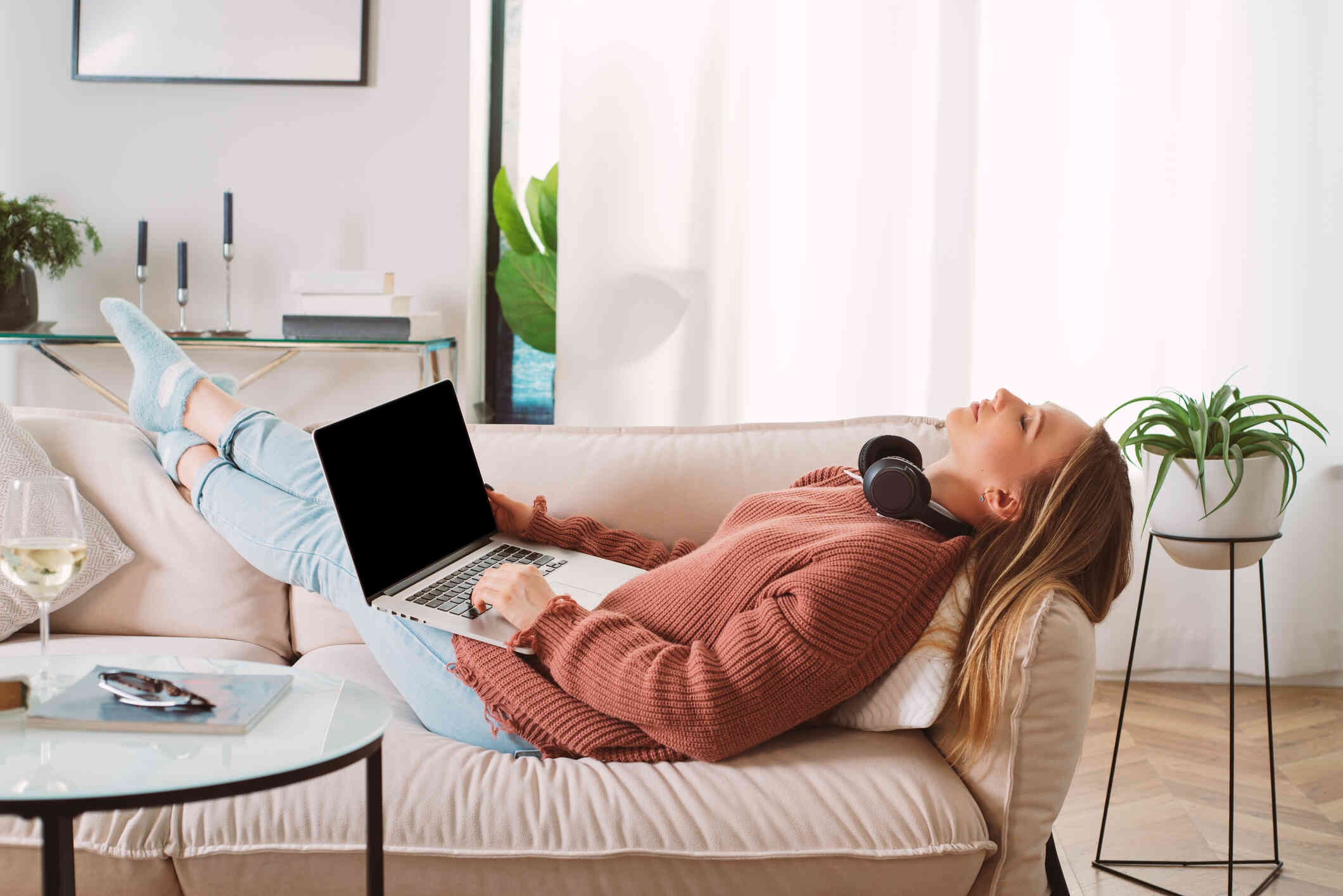 A woman in a red sweater falls asleep while reclining on the couch with her laptop still open in her lap and headphones around her neck.