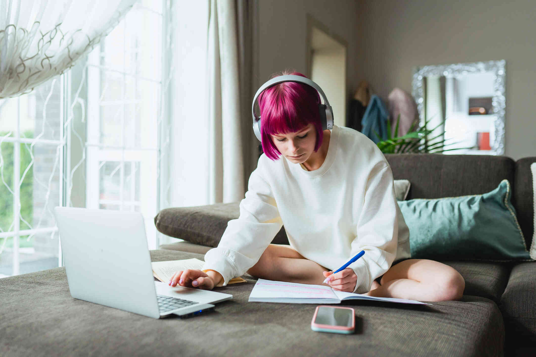 A young woman with pink hair and a white sweatshirt sits on her couch with a laptop while writing in a notebook with a focused expression.