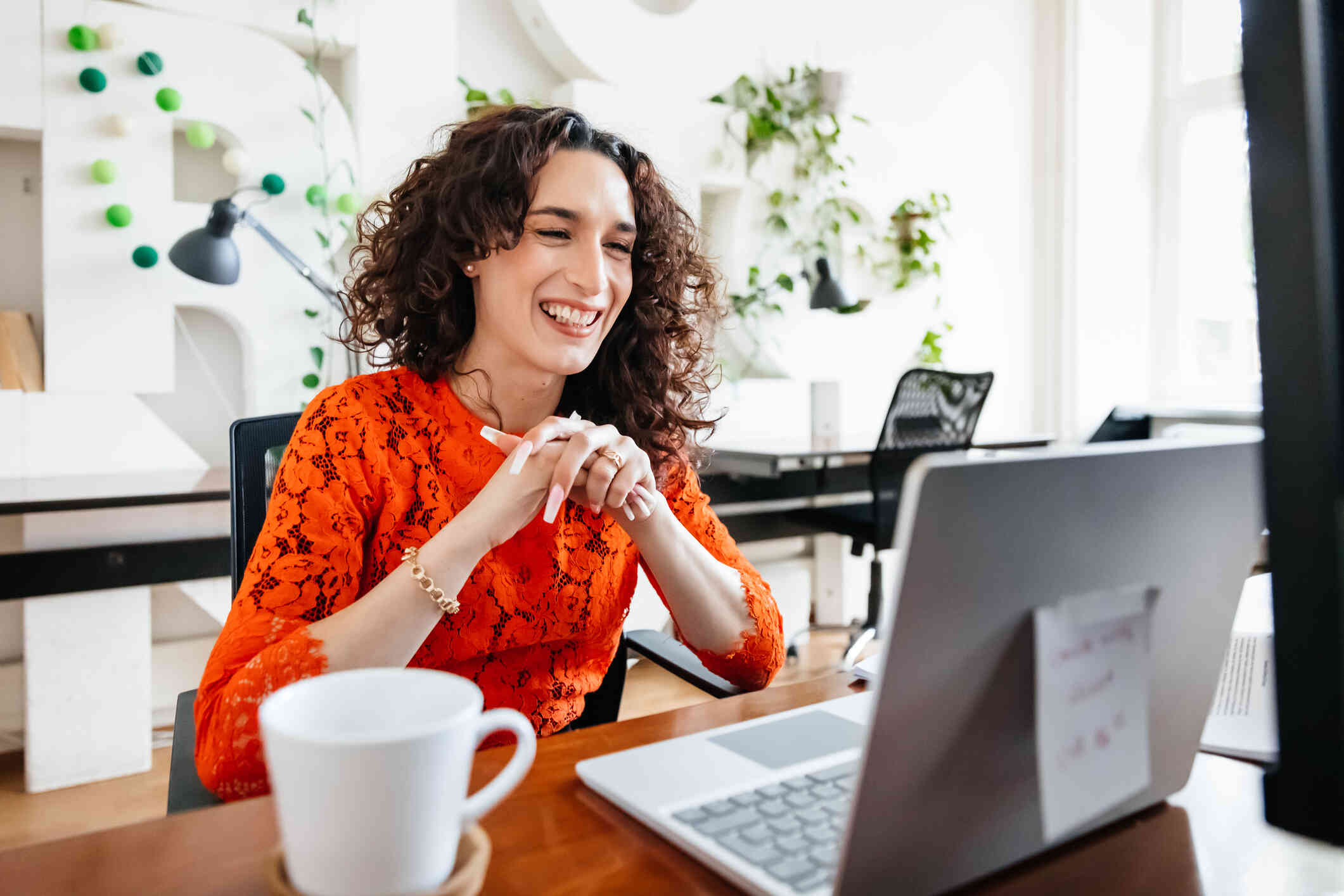 A woman seated at a table smiles at her laptop screen, appearing to be in a video call.