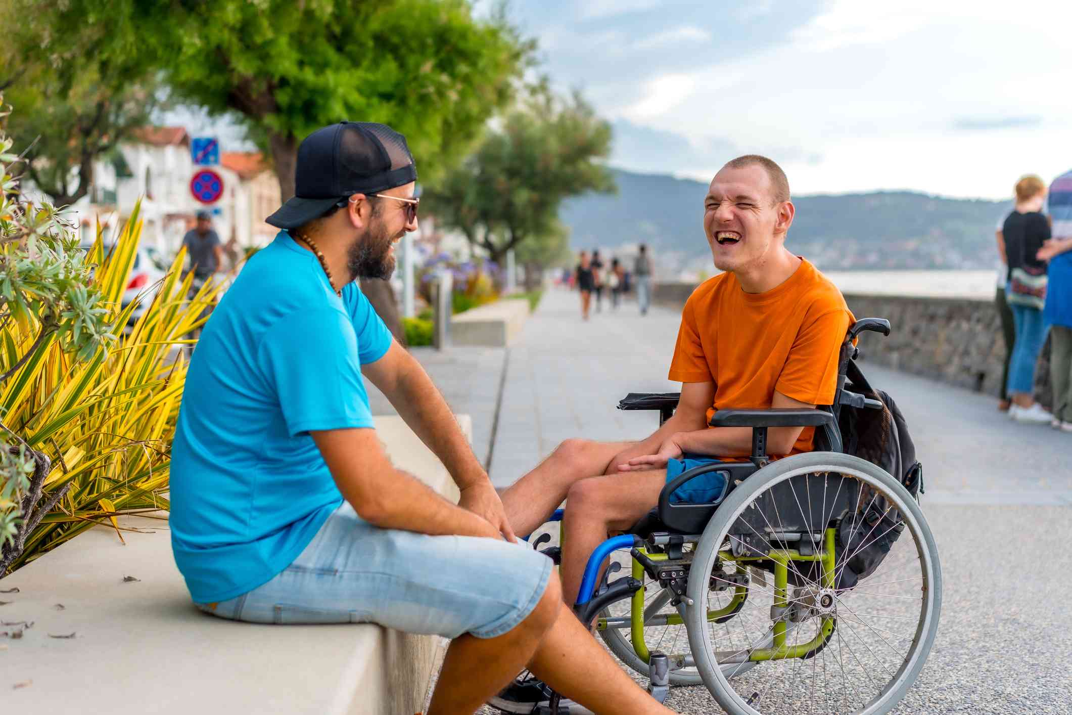 A man in a wheelchair and his male friend in a blue shirt sits outside together on a sunny day while chatting and smiling.