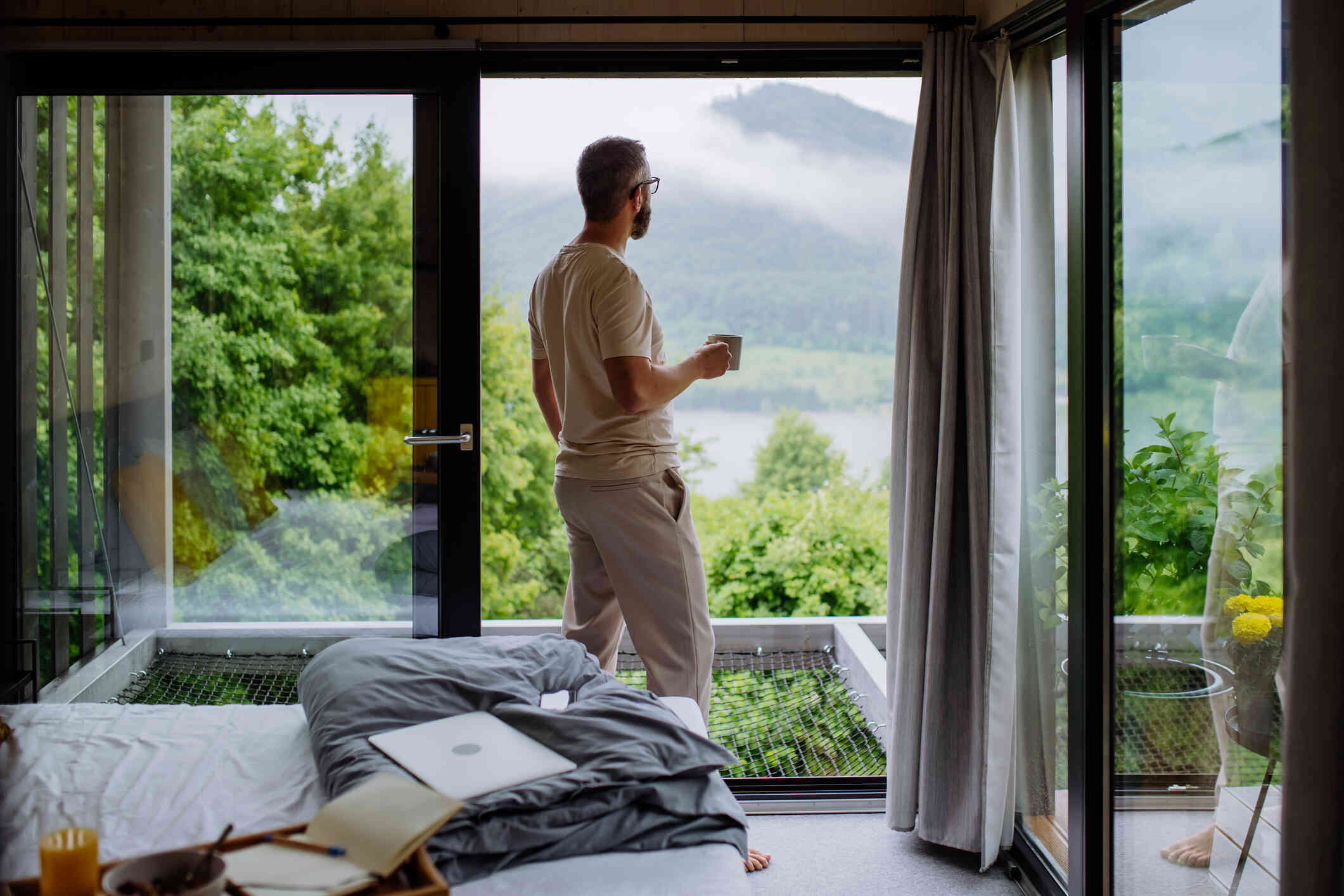 A man with glasses gazes out of  a window in his bedroom while holding a cup of coffee.