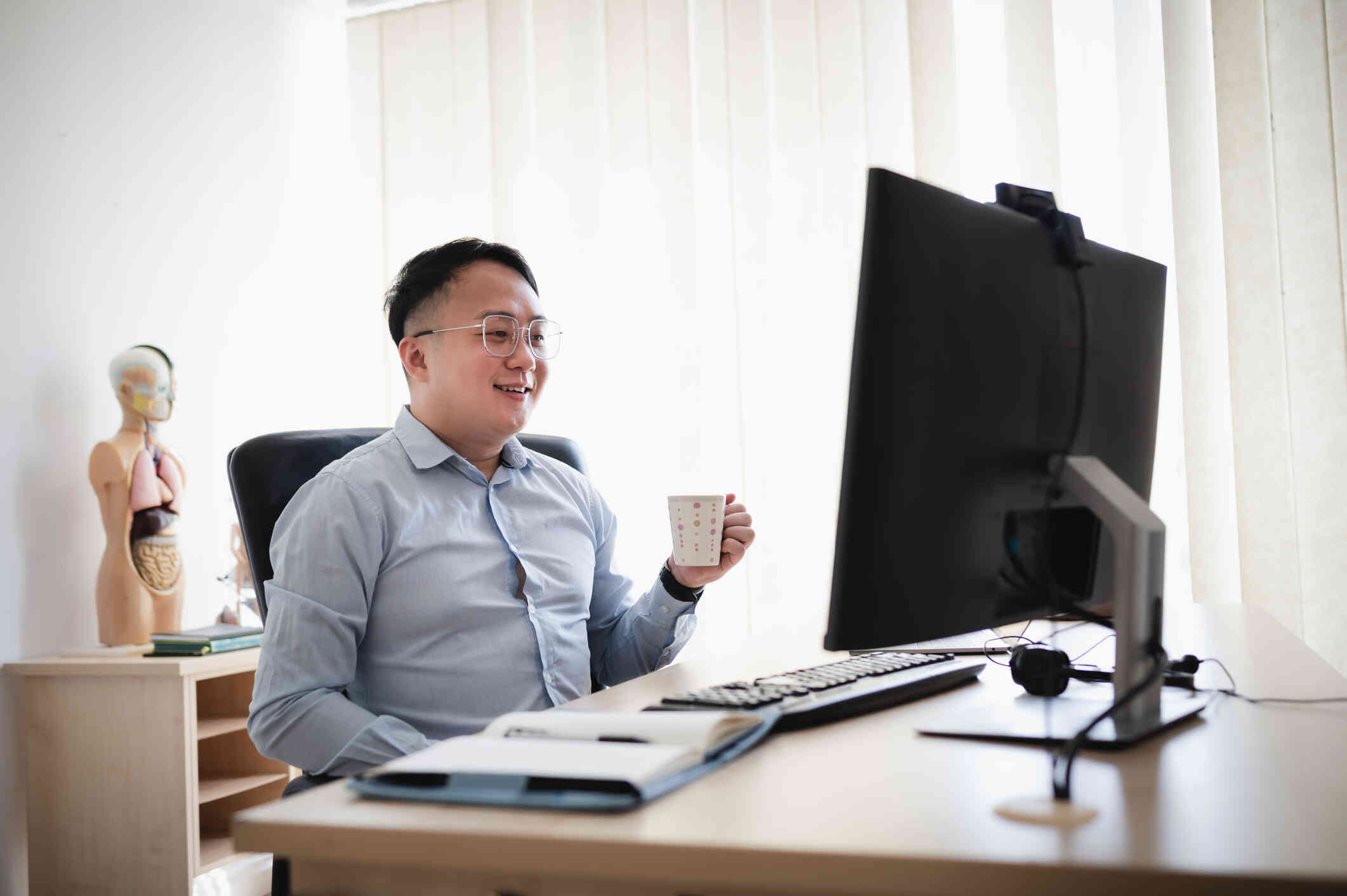 A man smiles at his laptop screen while holding a cup of coffee.