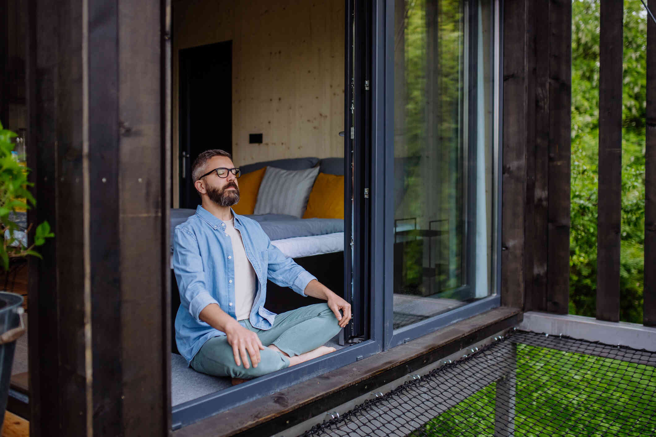 A man in a blue shirt with glasses sits on the ground outside while sitting in a yoga pose.