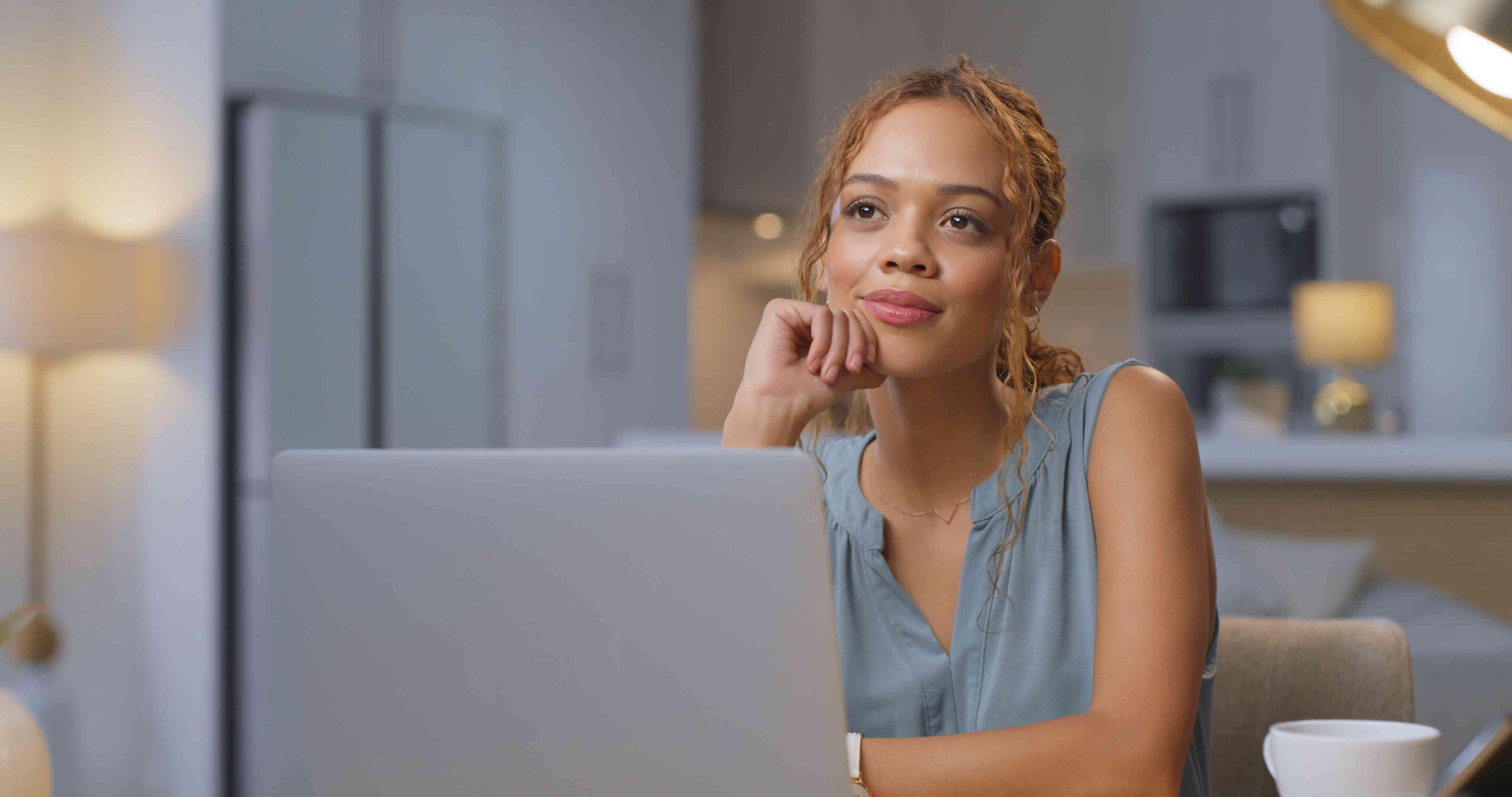 A woman in a blue blouse sits at her table in her home with her laptop open infront of her as she gazes off while deep in thought with a soft smile.