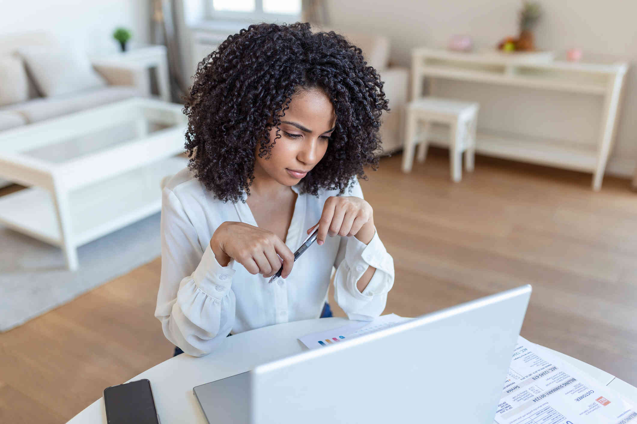 Awoman in a white button down shirt sits at her computer desk and loks at the screen while holding a pen in her hand.