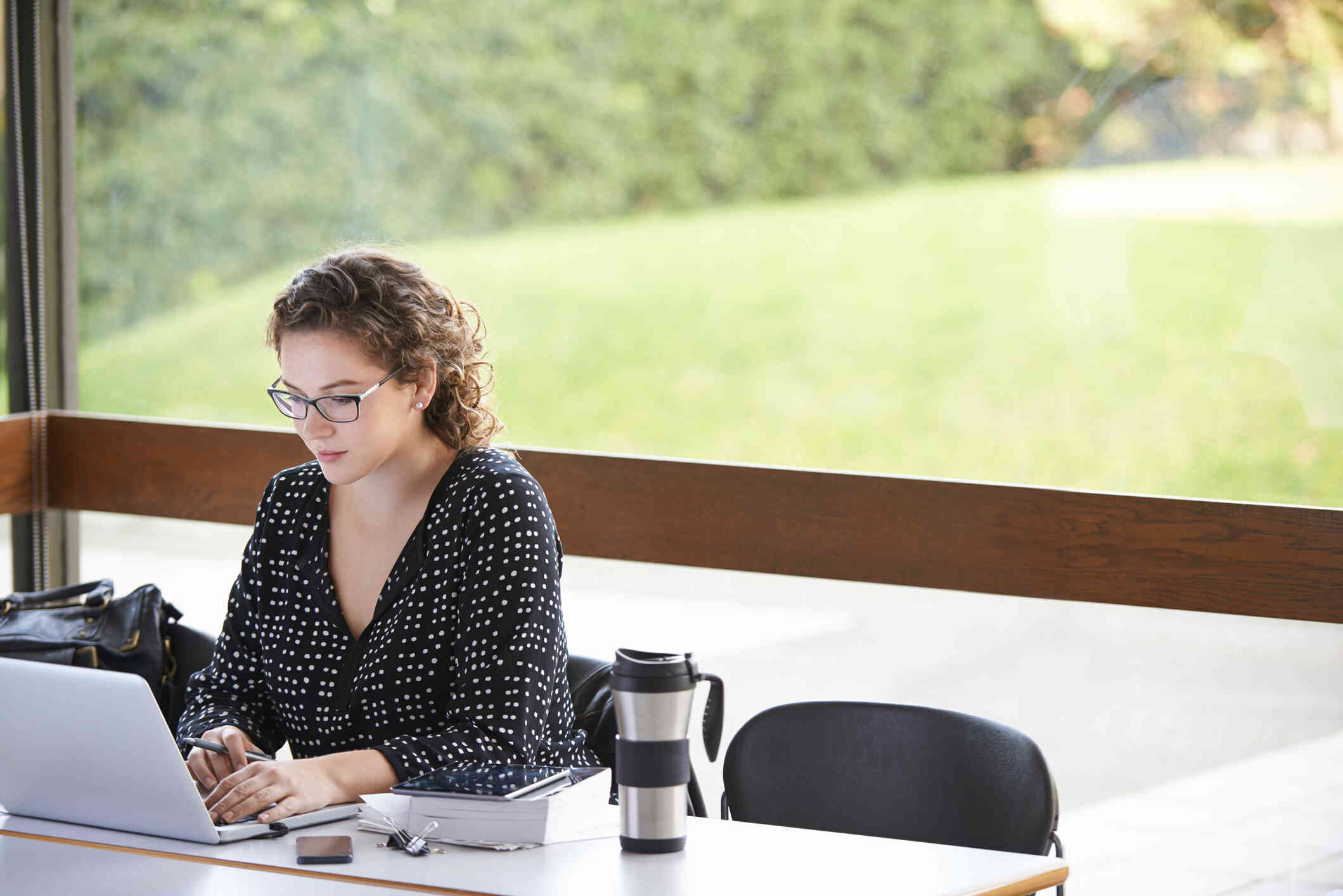 A woman sits at her desk, working on her laptop with an outdoor view through the windows.